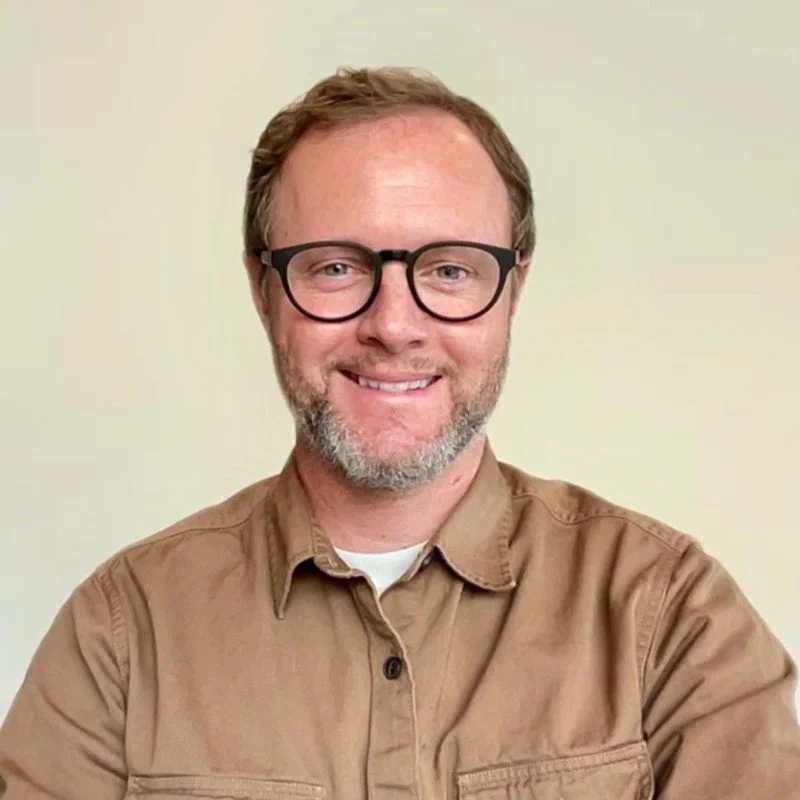 A person wearing glasses and a brown shirt, smiling at the camera against a plain background.