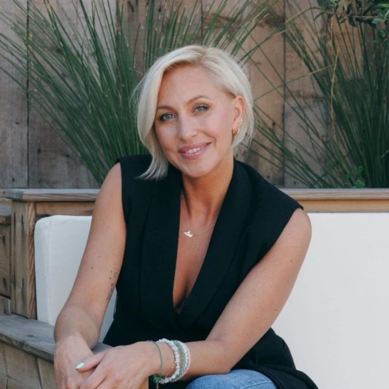 Woman with short blonde hair sitting on a cushion, wearing a black sleeveless top and smiling, with greenery in the background.