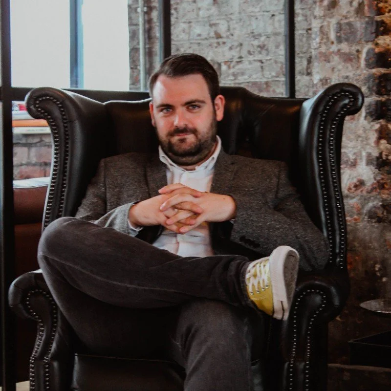 Man sitting in a leather armchair with a gray jacket, white shirt, and yellow sneakers, in an industrial-style room.