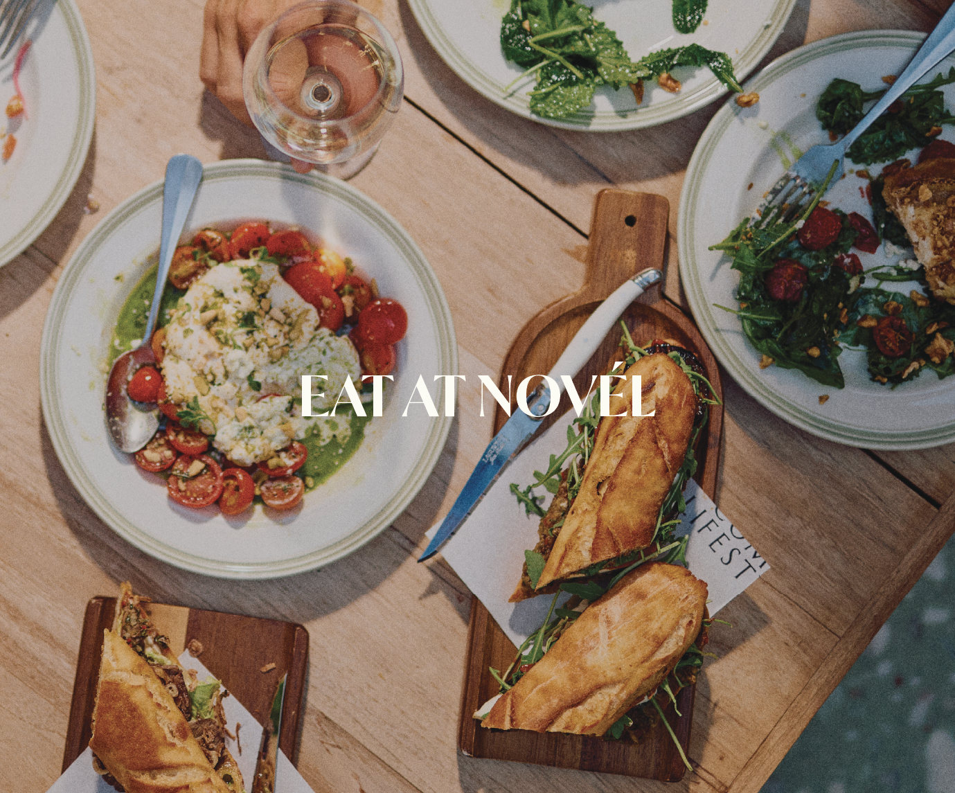 A variety of dishes on a wooden table, including salads, sandwiches, and wine, with the text 'EAT AT NOVEL' overlayed.
