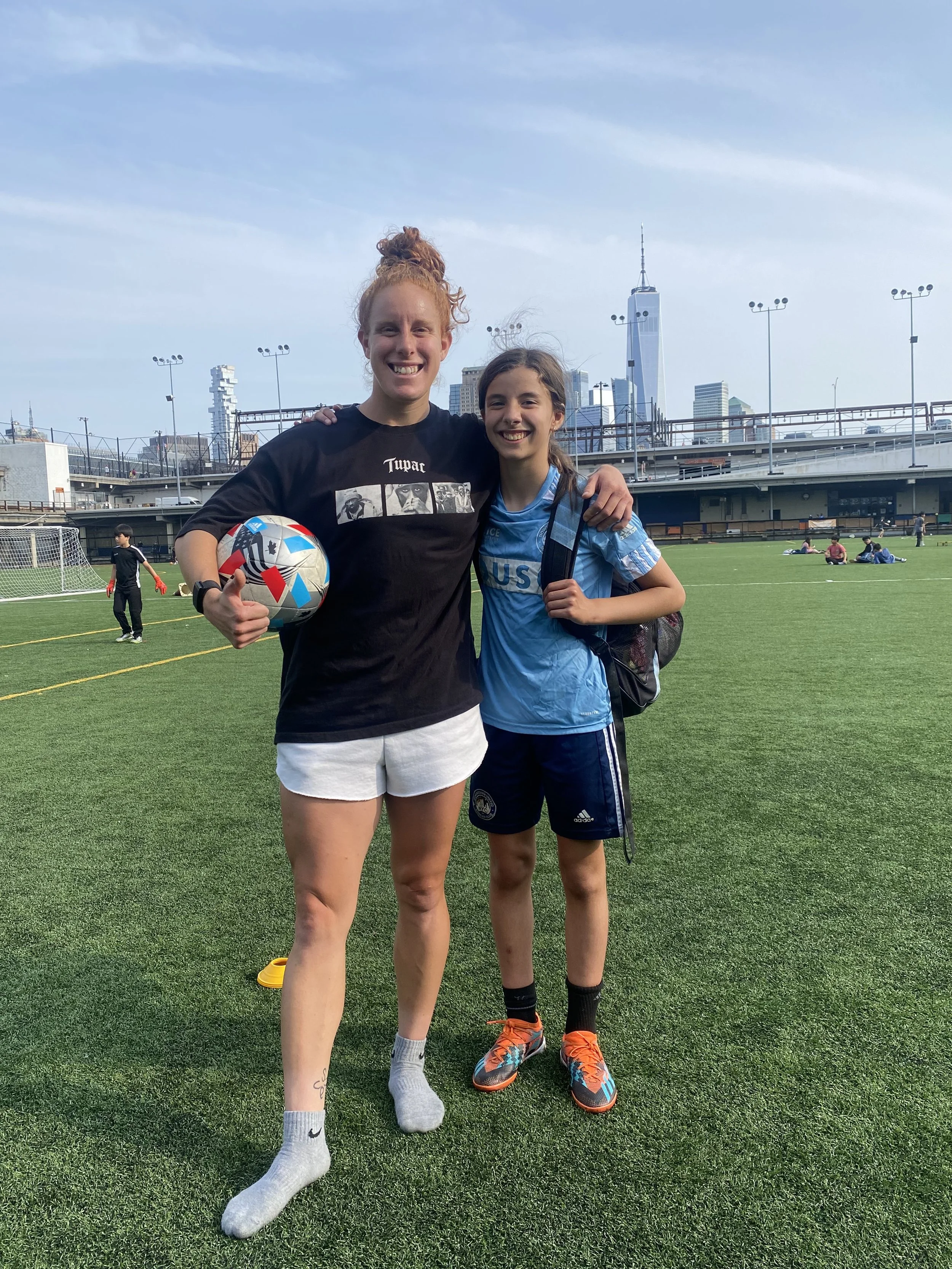 Two young women smiling on a soccer field with city buildings in the background, one holding a soccer ball.