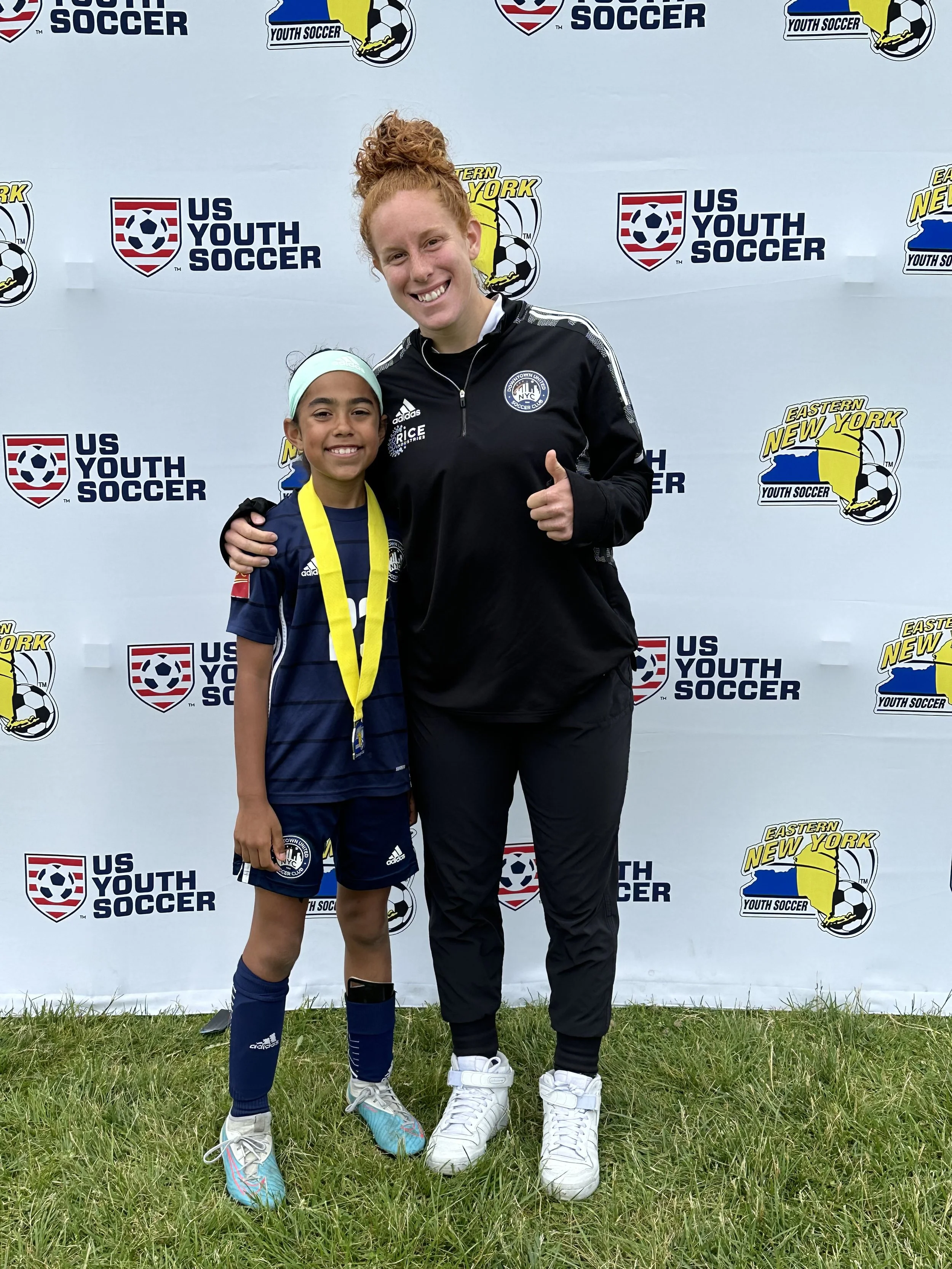 A young girl in a blue soccer uniform with a medal around her neck standing with an adult in black sportswear in front of a Disney backdrop at a youth soccer event. The adult is giving a thumbs-up and they are both smiling. The backdrop features multiple logos for US Youth Soccer and Eastern New York Youth Soccer.