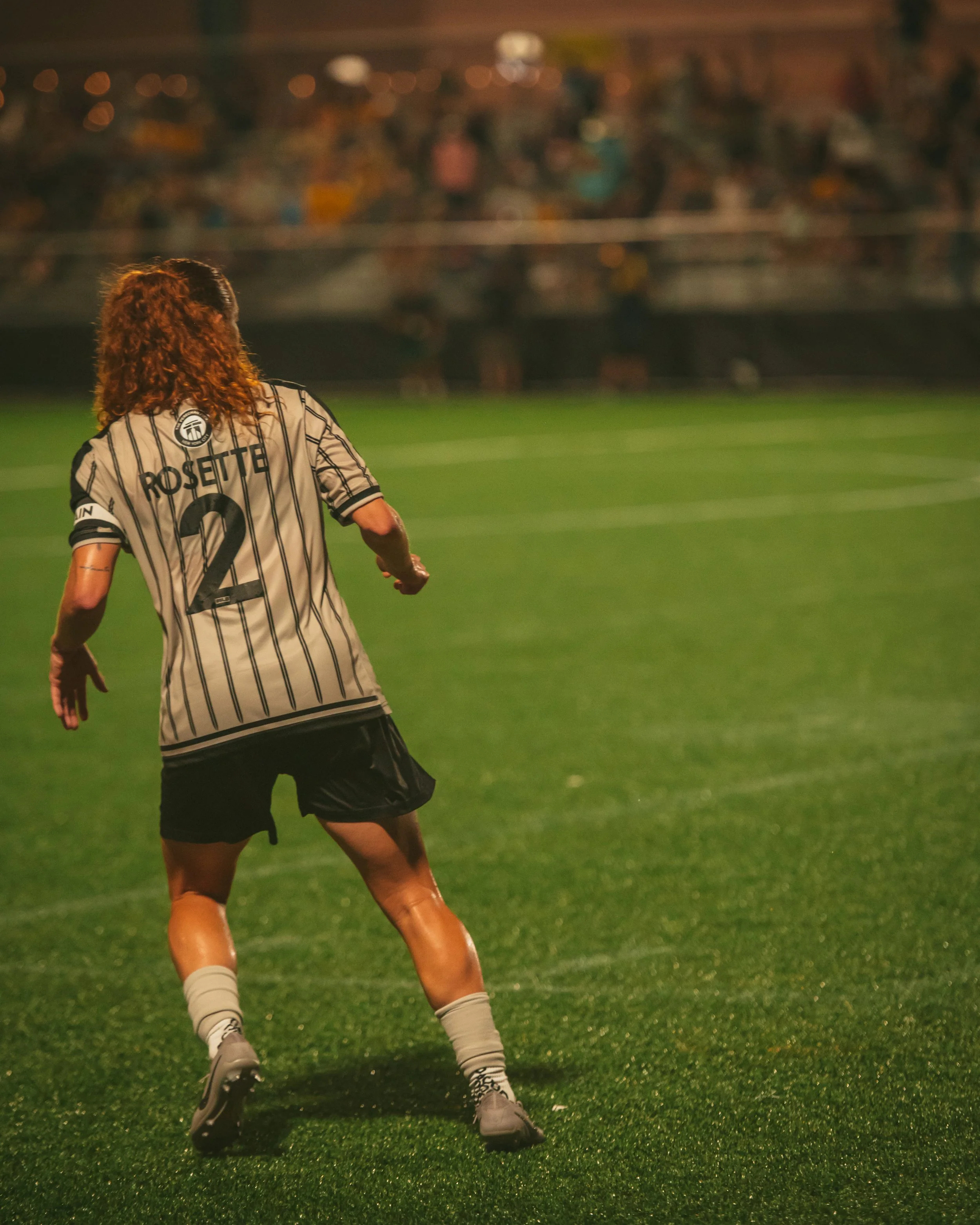 A female soccer player with curly red hair on a green field during night, wearing a black and white striped jersey with the name 'Rosette' and the number 2, black shorts, white socks, and gray cleats.