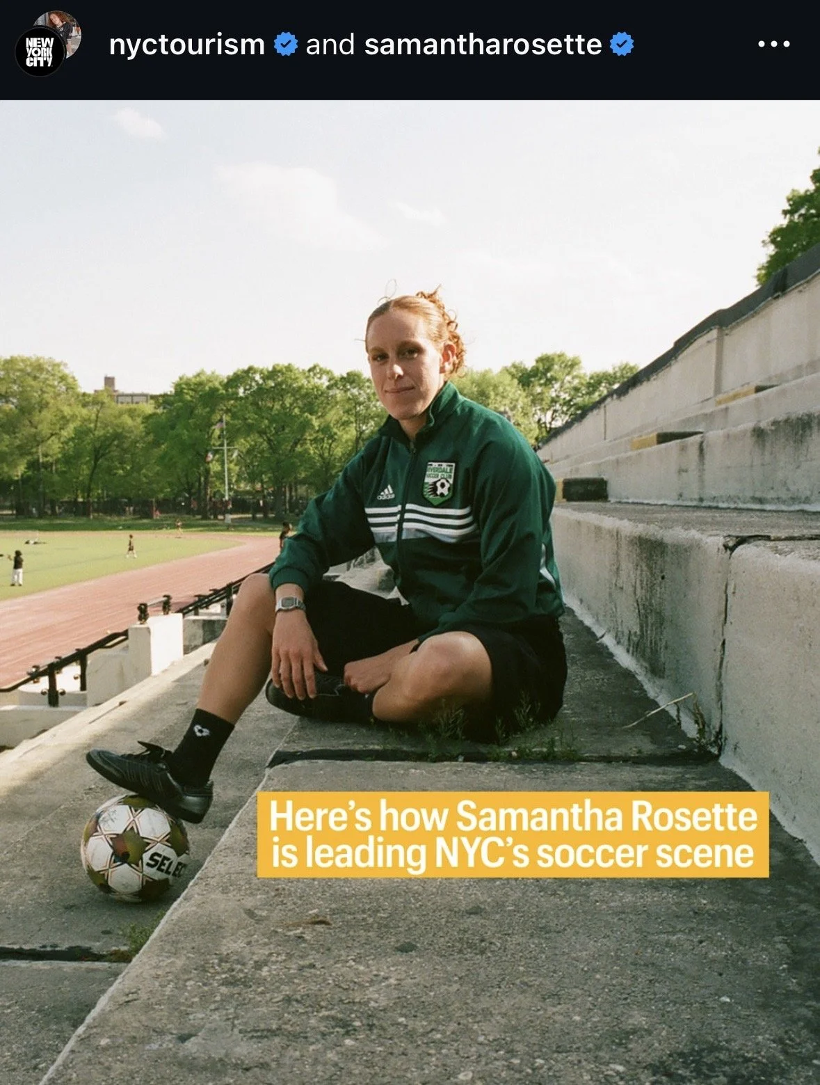 A young woman with red hair sitting on stadium bleachers with one foot on a soccer ball, wearing a green NY soccer team jacket, black shorts, socks, and sneakers, at an outdoor track and soccer field.