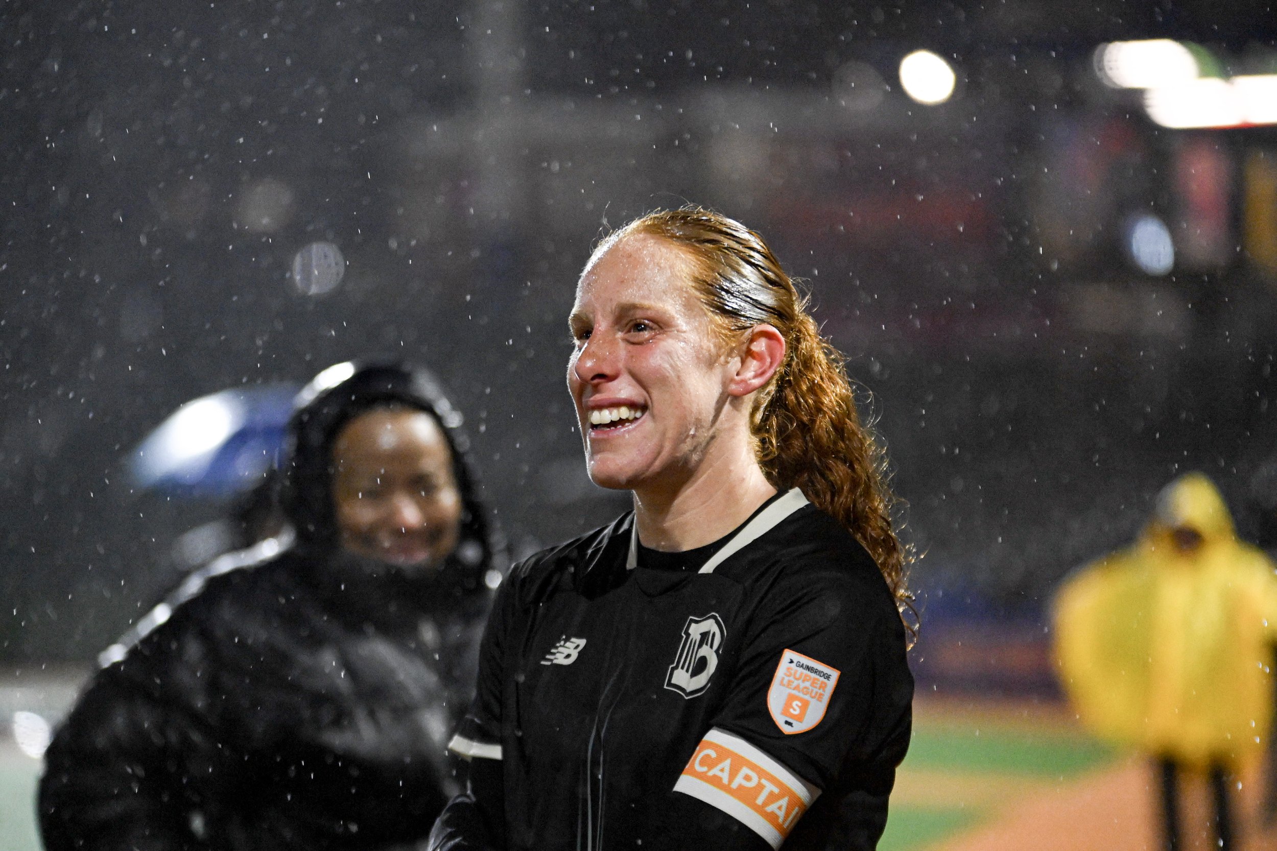 A woman with red hair and a black sports jersey smiling during a rainstorm at a sports event. She is wearing a captain's armband on her right arm and is surrounded by other people in athletic gear.