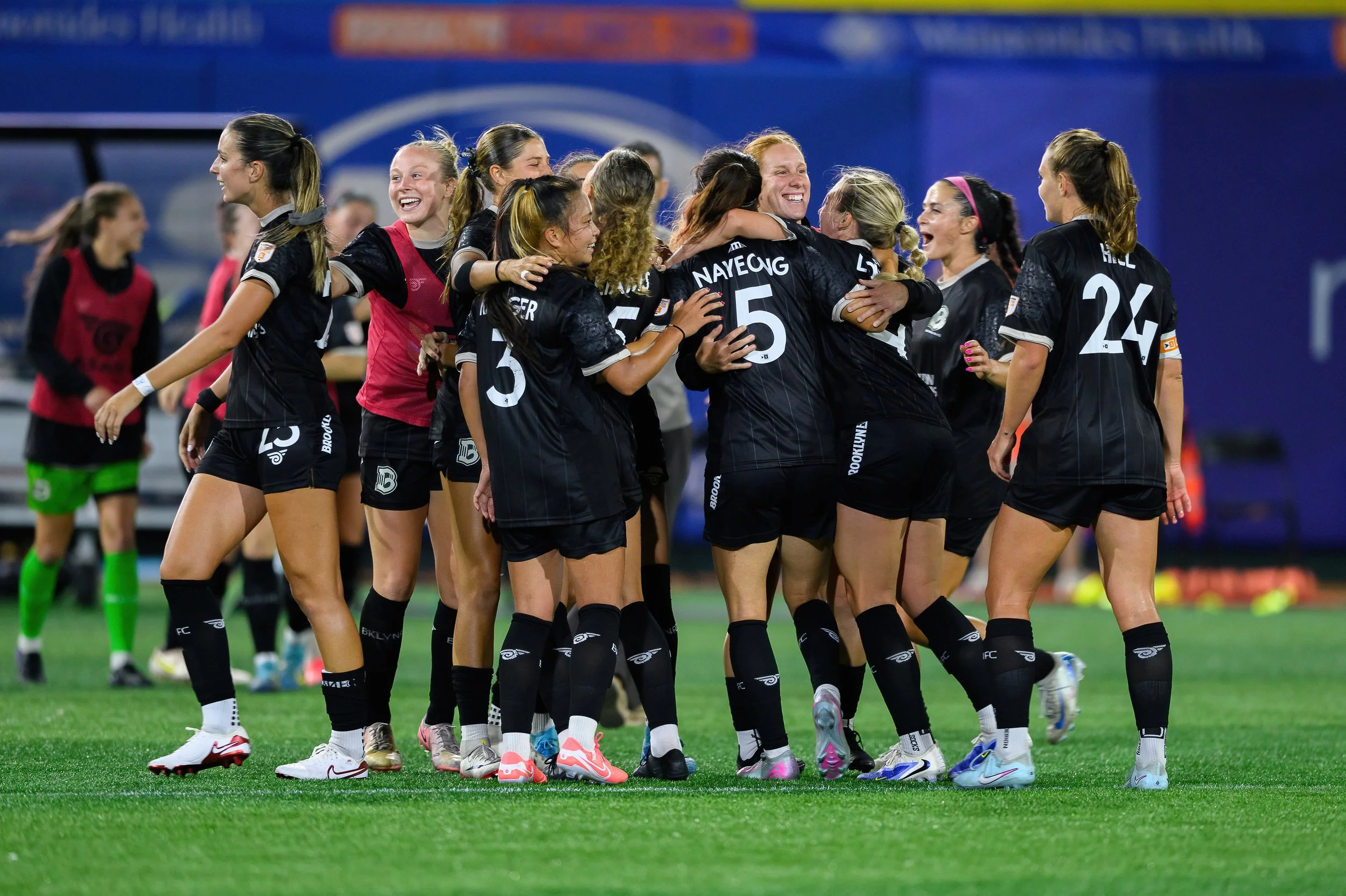 A group of female soccer players celebrating on the field, hugging and smiling.
