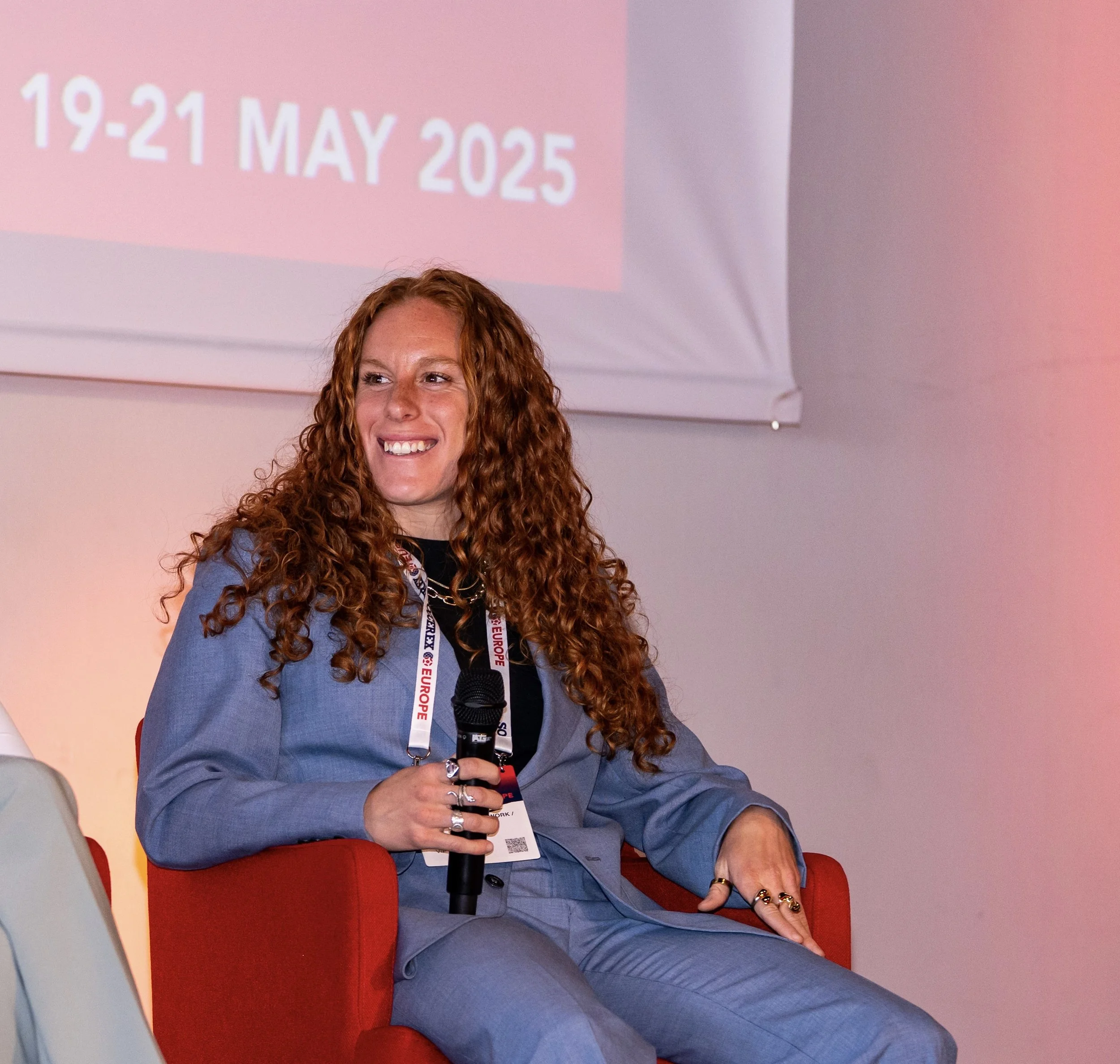 A woman with curly red hair sitting on a red chair, smiling, holding a microphone, wearing a blue blazer, a black shirt, and a lanyard with event tags, against a wall with a projection displaying dates in May 2025.