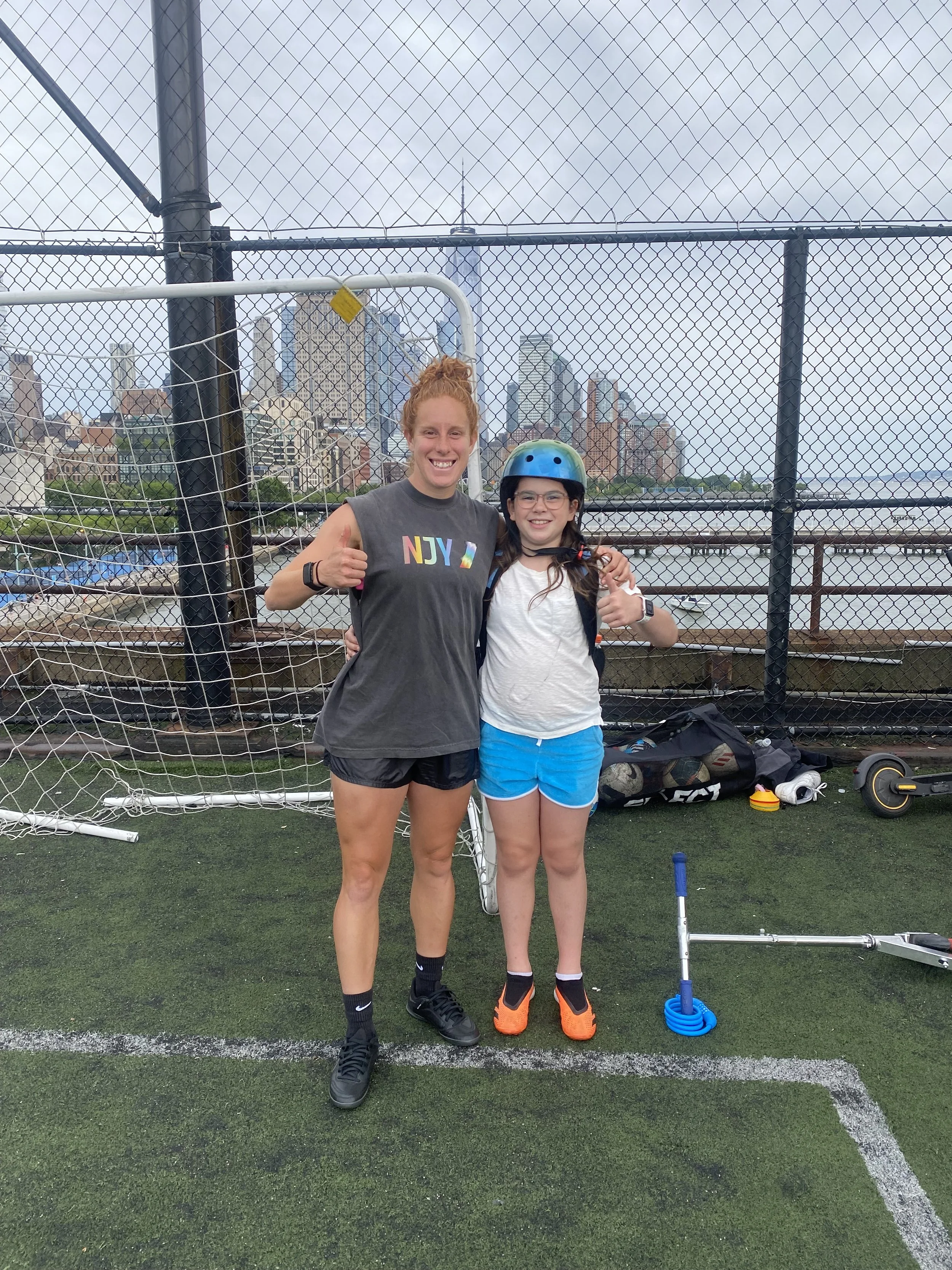 Two young women stand on a sports field in front of a fence with a city skyline and water in the background. One woman has red hair in a bun and is giving a thumbs-up, wearing a sleeveless gray shirt with colorful letters and black shorts. The other woman has long brown hair, glasses, a helmet, a white shirt, and blue shorts. Both are smiling and appear to have finished a sports activity.