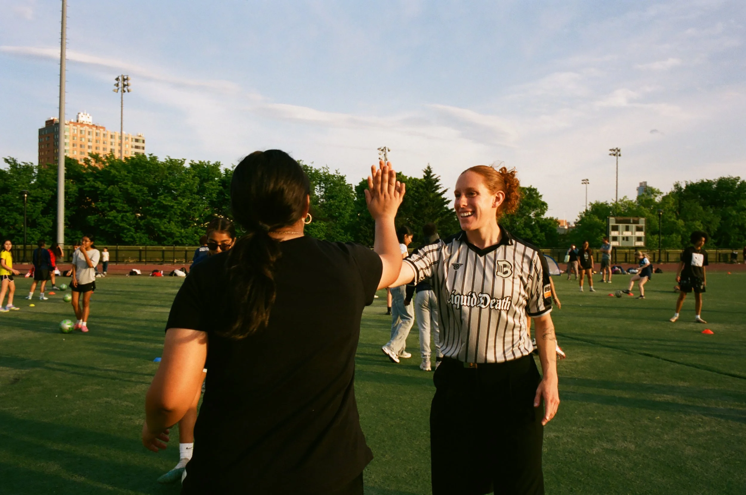 Two women high-fiving on a sports field with people playing soccer in the background.