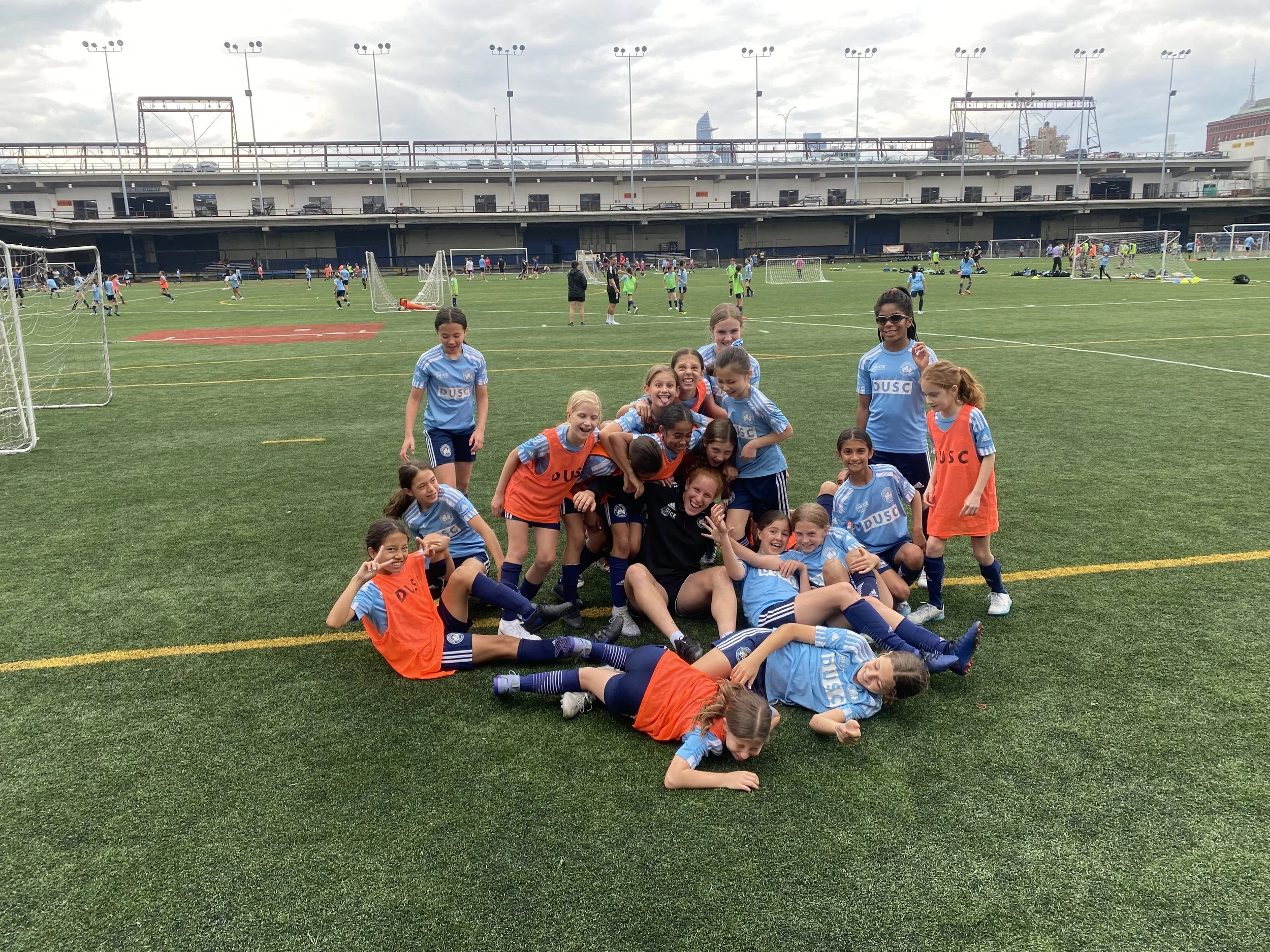 Group of young girls in soccer uniforms, some with orange vests, are posing for a photo on a soccer field. There are many children playing soccer and practicing in the background, with a stadium structure, cityscape, and overcast sky above.