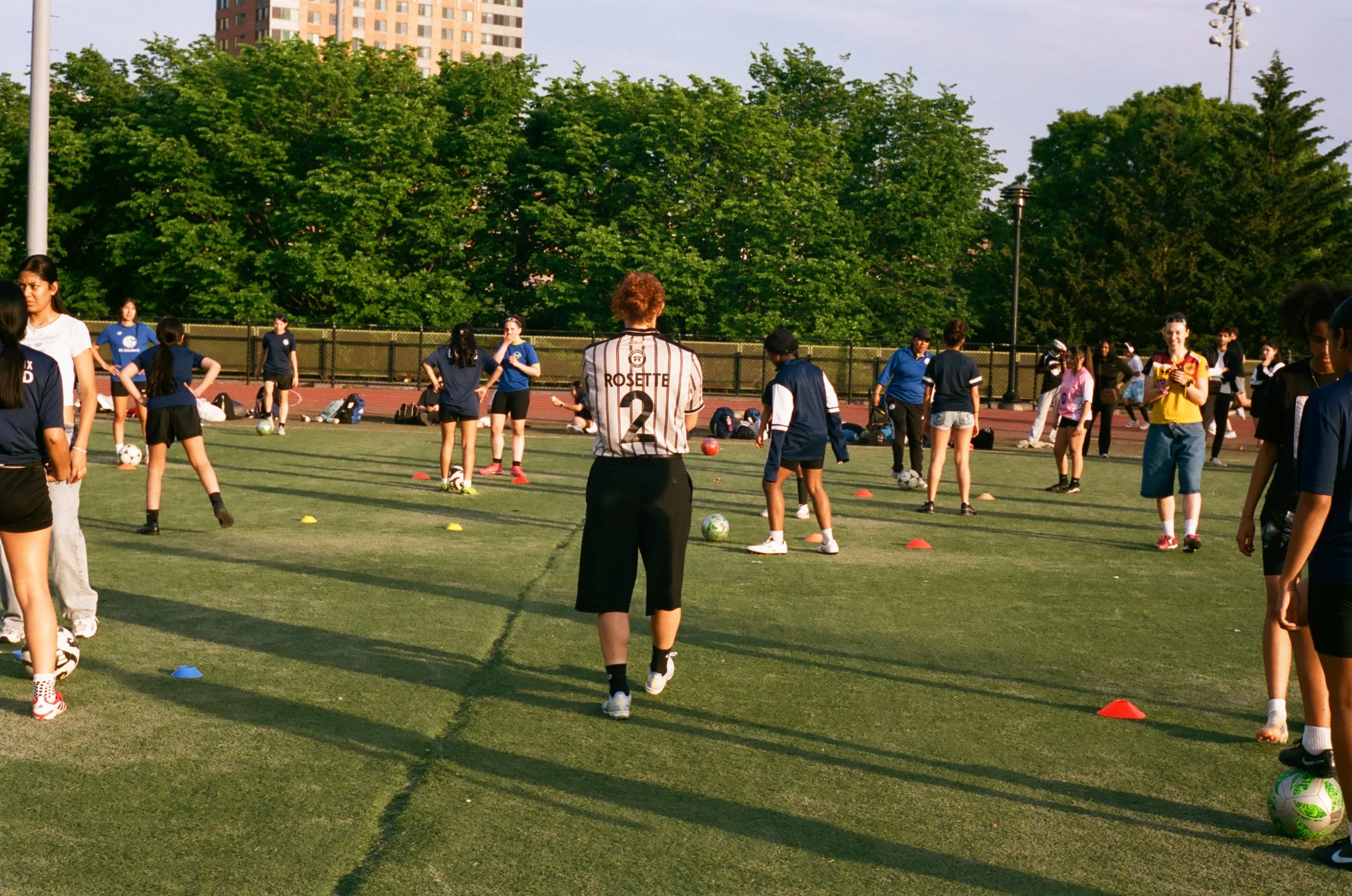 Women and girls participating in a soccer training session outdoors, with soccer balls and orange cones on the grass field, surrounded by green trees and tall buildings in the background.