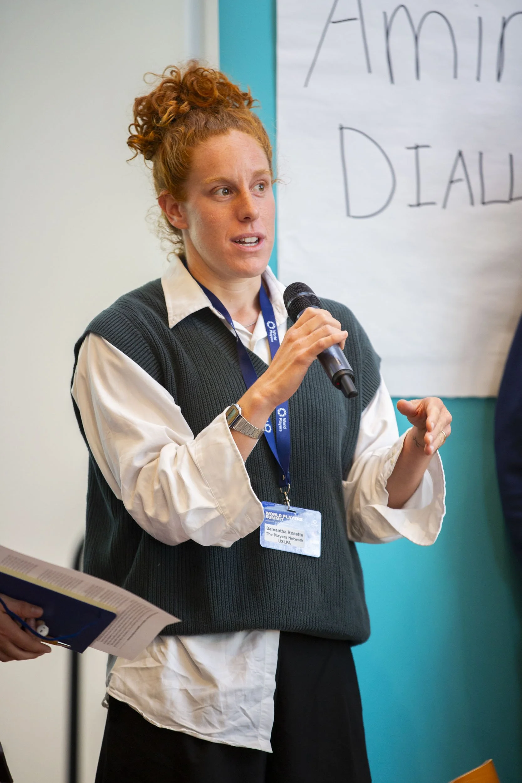 A woman with curly red hair holding a microphone and wearing a lanyard with a badge presents in a classroom or conference room.