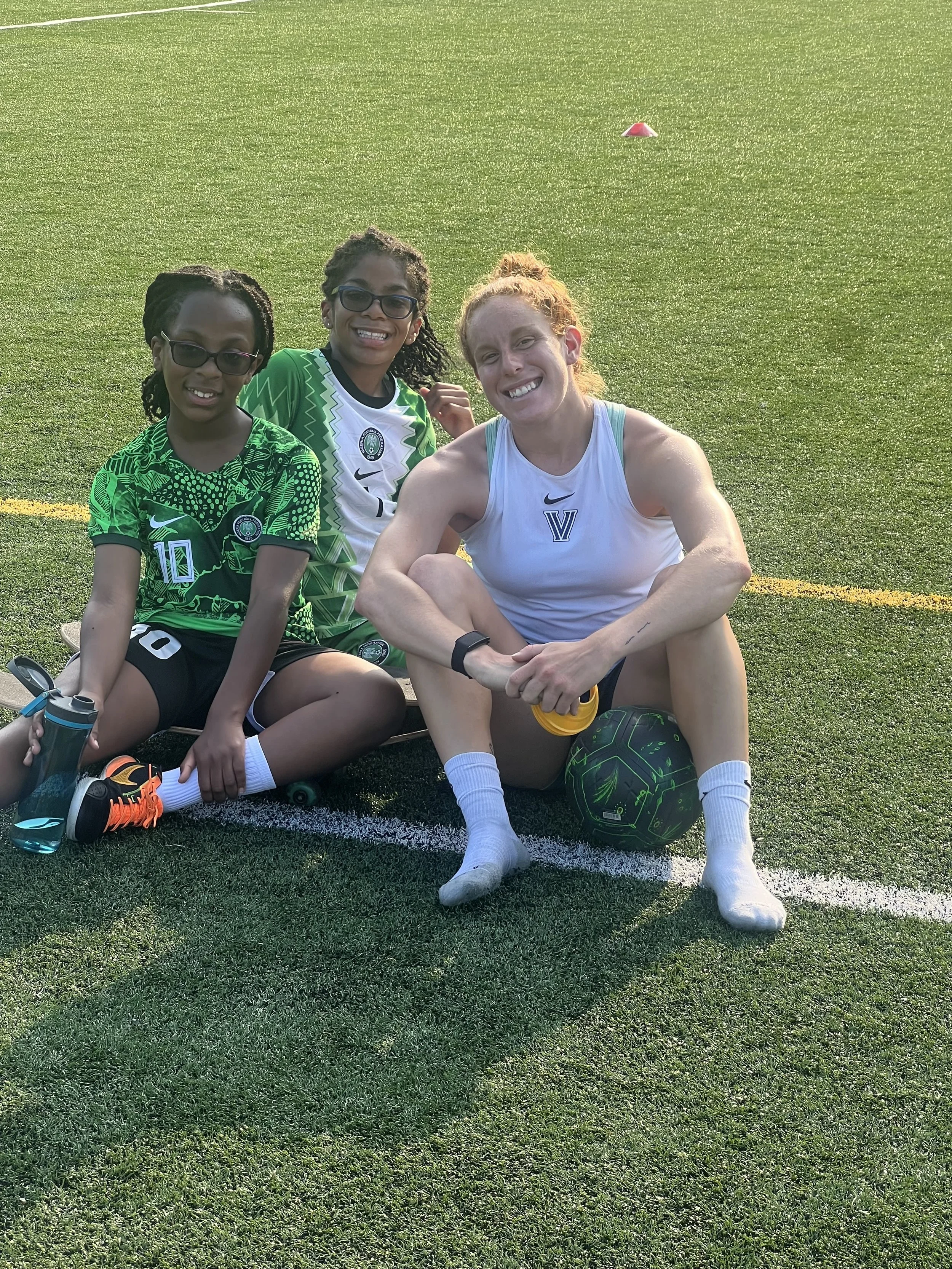 Three women sitting on a football field with a green grass surface and a small orange cone in the background, smiling.
