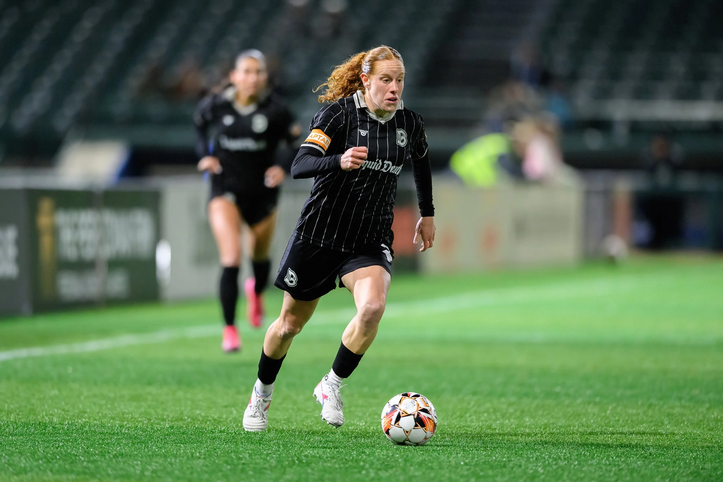 A woman playing soccer on a green field, wearing a black striped jersey with a captain's armband, controlling a soccer ball, with another player in the background.