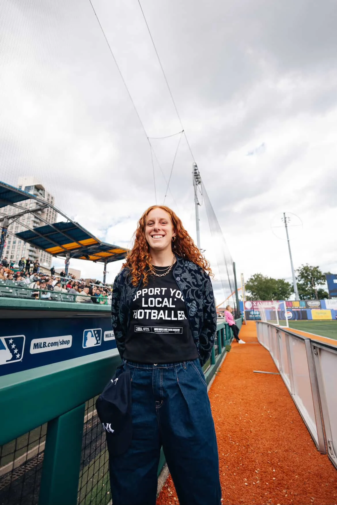 A woman with long curly red hair smiling at a baseball stadium, wearing a black T-shirt that says 'Support Your Local Footballer' and baggy blue pants, standing near the fence on a cloudy day.