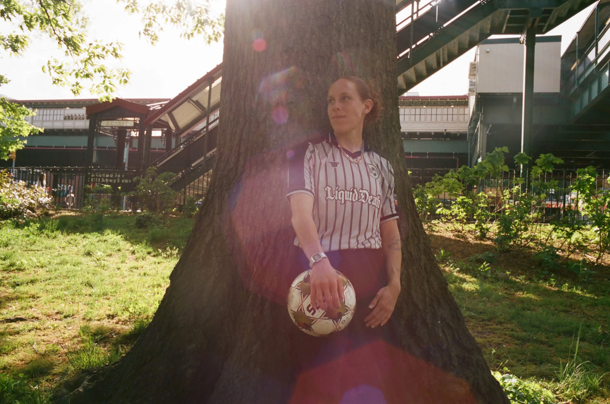 Person with short red hair wearing a black and white striped sports jersey, holding a soccer ball, leaning against a large tree in a park, with a subway station in the background.