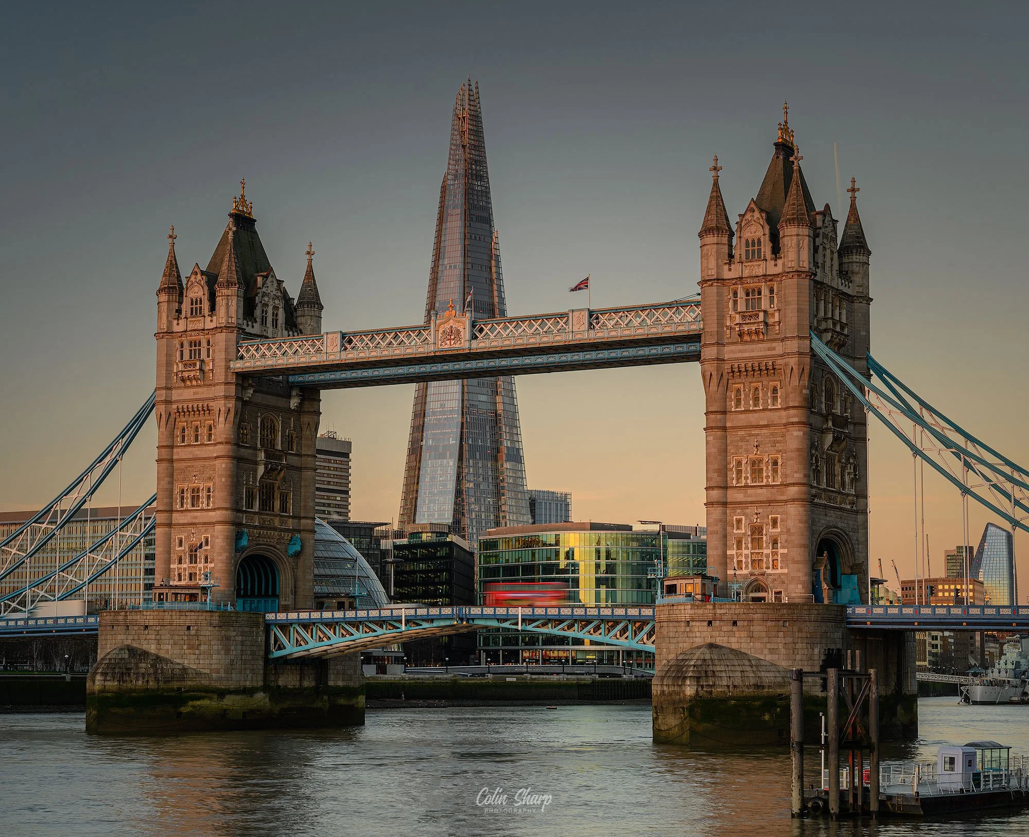 View of Tower Bridge in London during sunrise, with the Shard skyscraper in the background and calm water in the foreground.