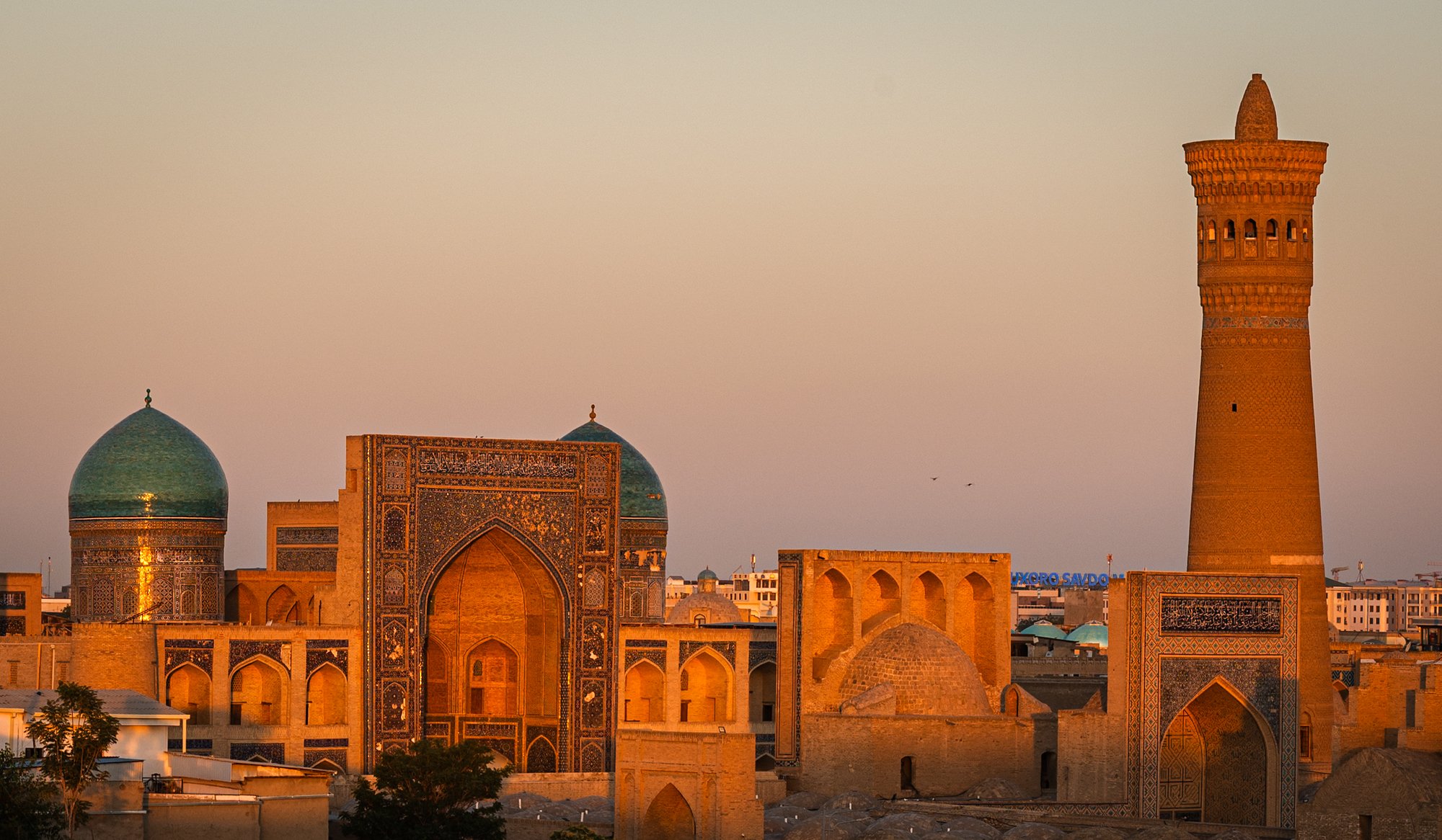 An image of the Bukhara Kalon Complex with the Kalon Minaret and Kalon Mosque and the Mir-i Arab Medressa photographed in the evening as the sun set with the buildings reflecting a warm orange light.