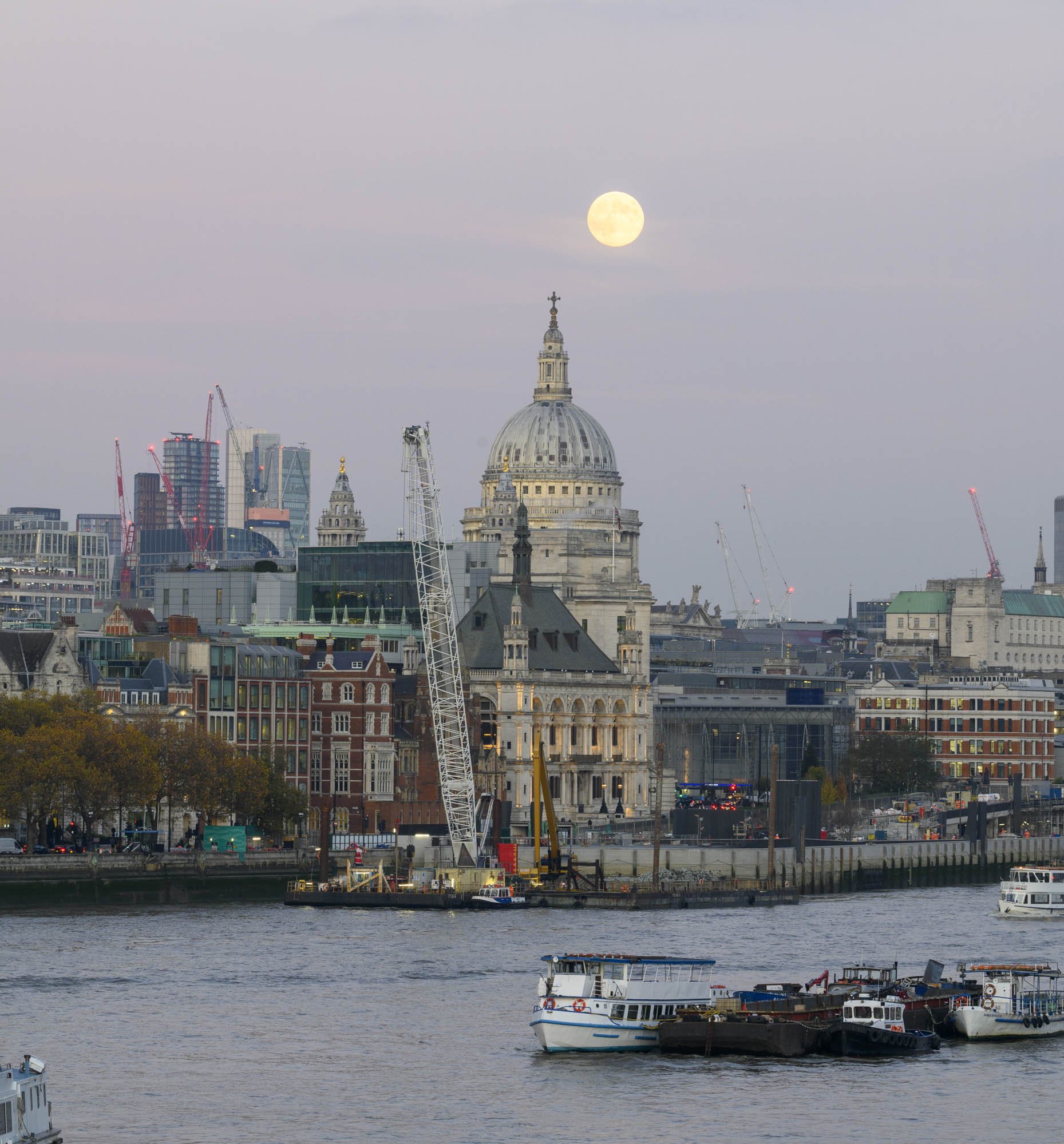 View of St. Paul's Cathedral in London with a full moon in the sky, boats on the river in the foreground, and a cityscape with construction cranes and modern buildings in the background.