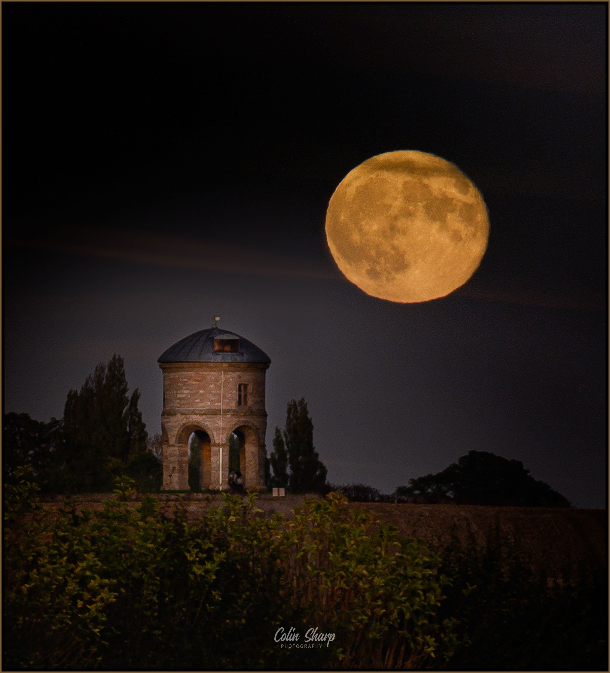 A full moon riding in the night sky above, Chesterton Windmill minus its sails that were being restored, a stone tower with a domed roof, surrounded by trees and bushes.