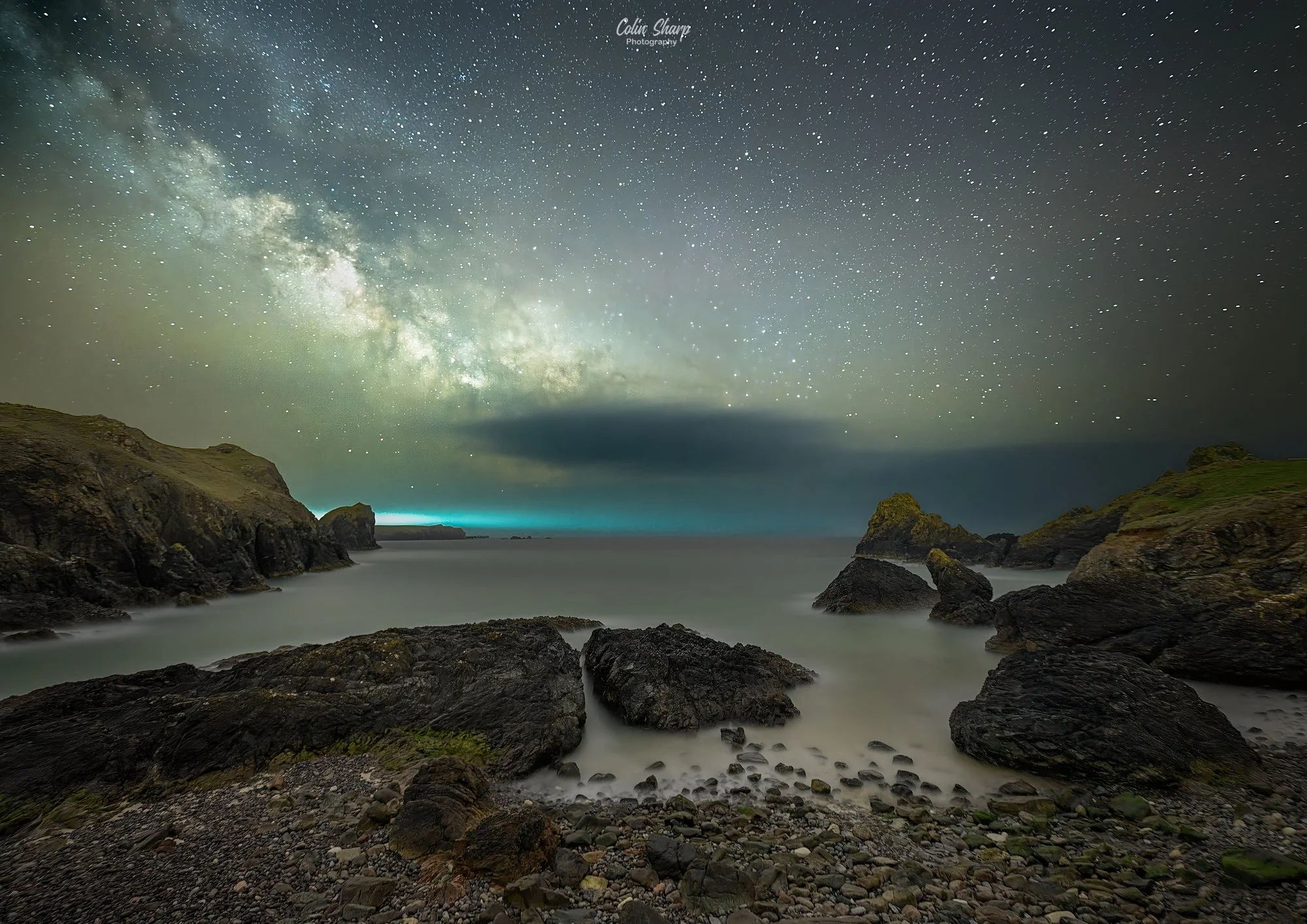 Night time coastal scene with starry sky and the Milky Way galaxy. Rocky shoreline with large rocks and pebble beach in the foreground. Cliff formations on both sides of the ocean, with a dark cloud hovering over the horizon.