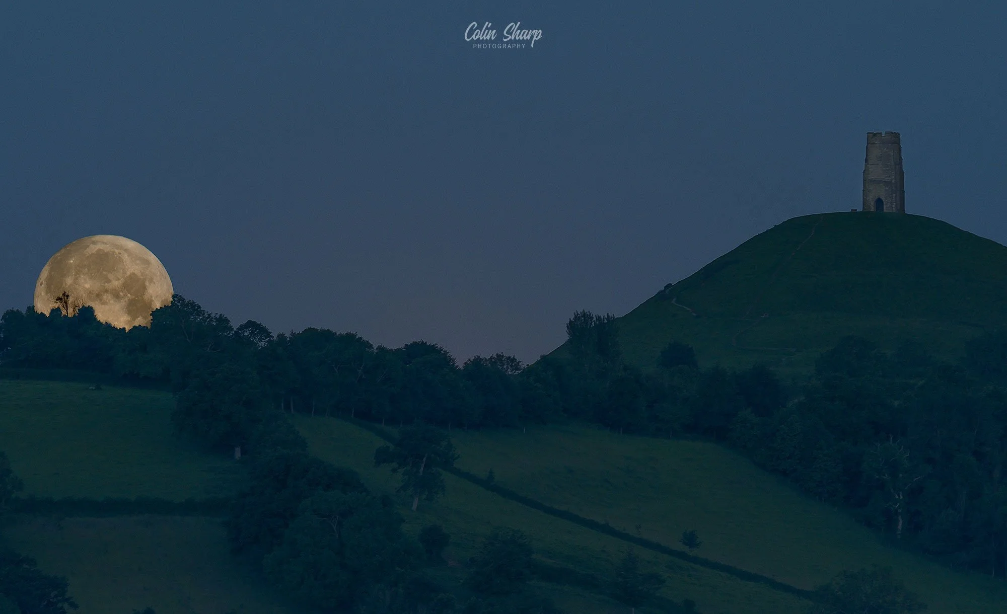 Nighttime landscape with rolling green hills, a stone tower called St Michaels Tower on Glastonbury Tor, on the right hill, trees in the foreground, and a large full Strawberry super moon near the horizon on the left.
