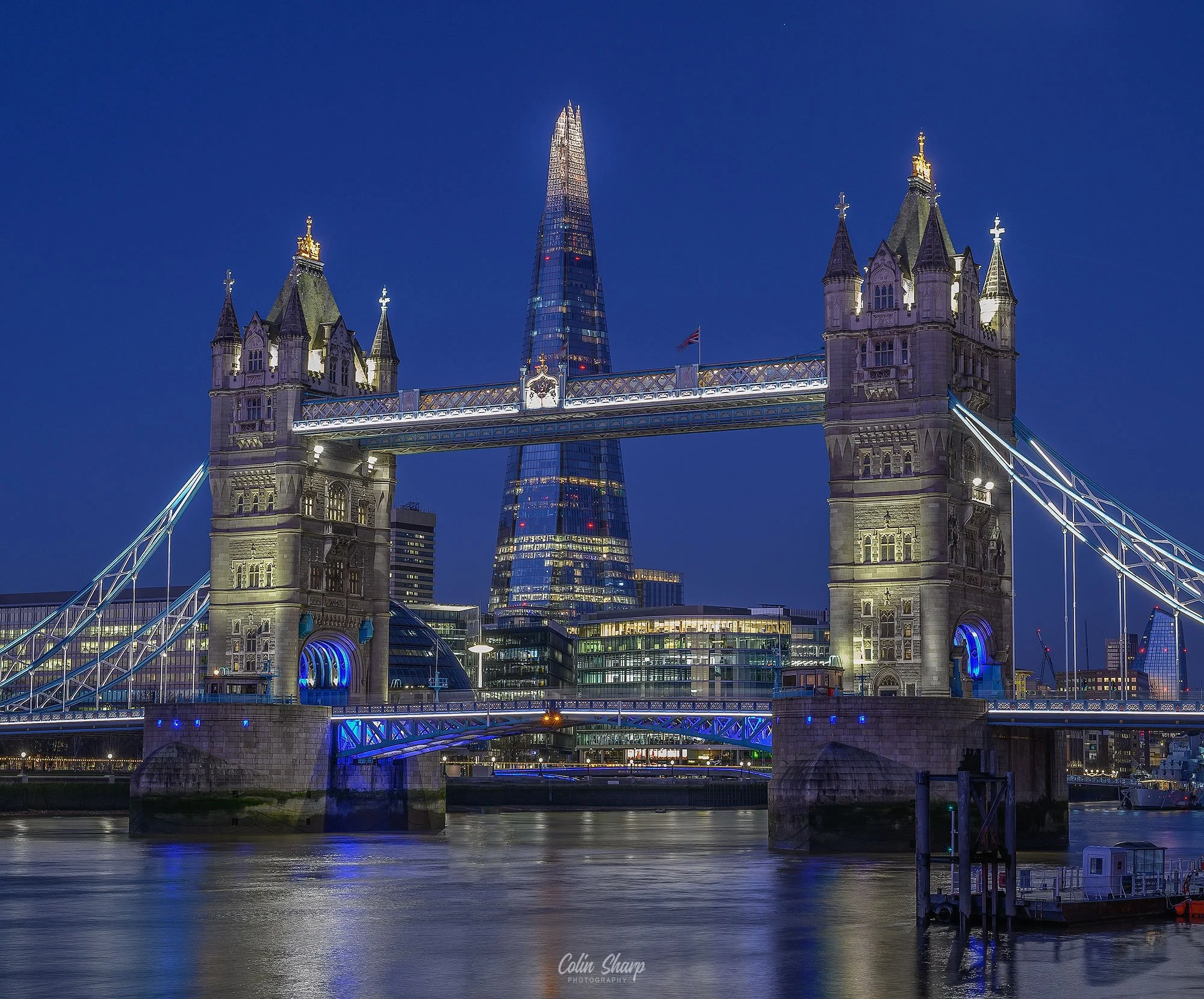 Blue Hour view of Tower Bridge in London with the Shard skyscraper in the background, illuminated and reflected on the River Thames.