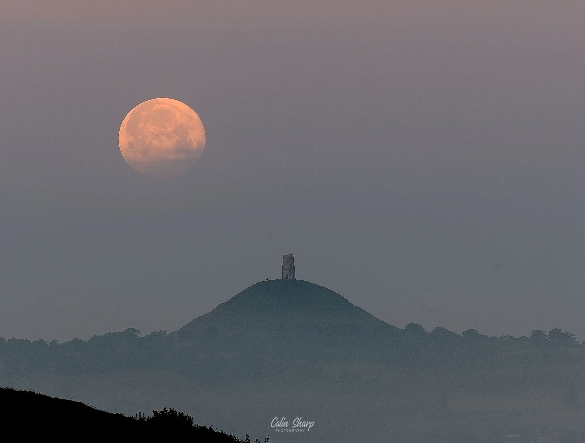 The Strawberry Super Moon setting behind a misty Glastonbury Tor and St Michaels Mount, an iconic tower on top of the Tor, with cloudy sky and tree silhouettes at the bottom.