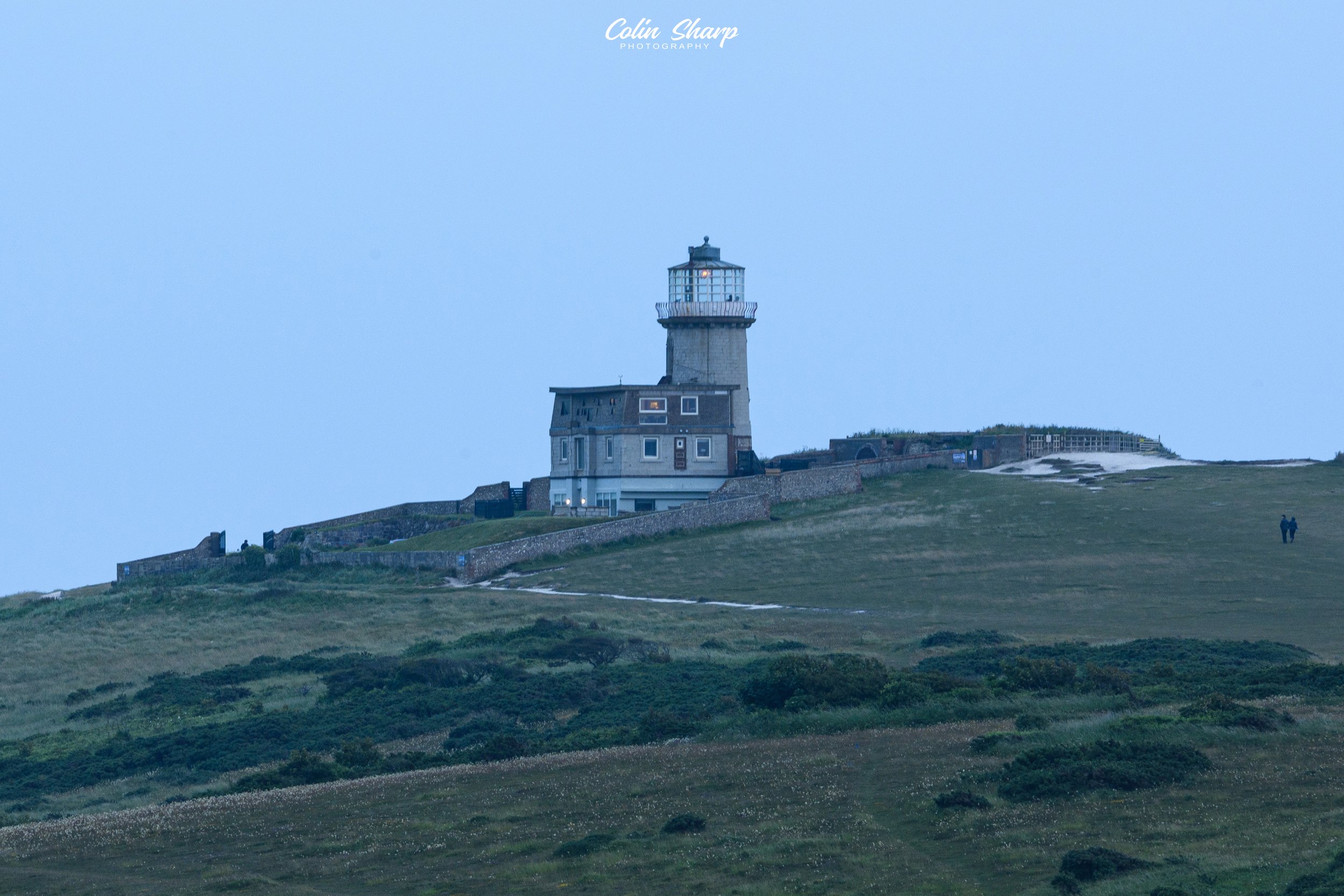 Belle Tout, in East Sussex, a lighthouse on a grassy hill with a few people walking nearby at dusk .