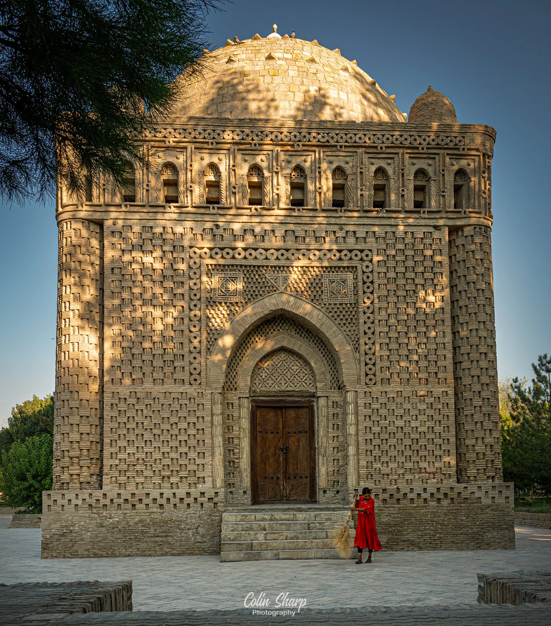 An image of the Ismail Samani Mausoleum in Bukhara, completed in 905 the town's oldest Islamic monument and taken during the morning golden hour whilst a woman dressed in red is sweeping the area in front of the entrance.