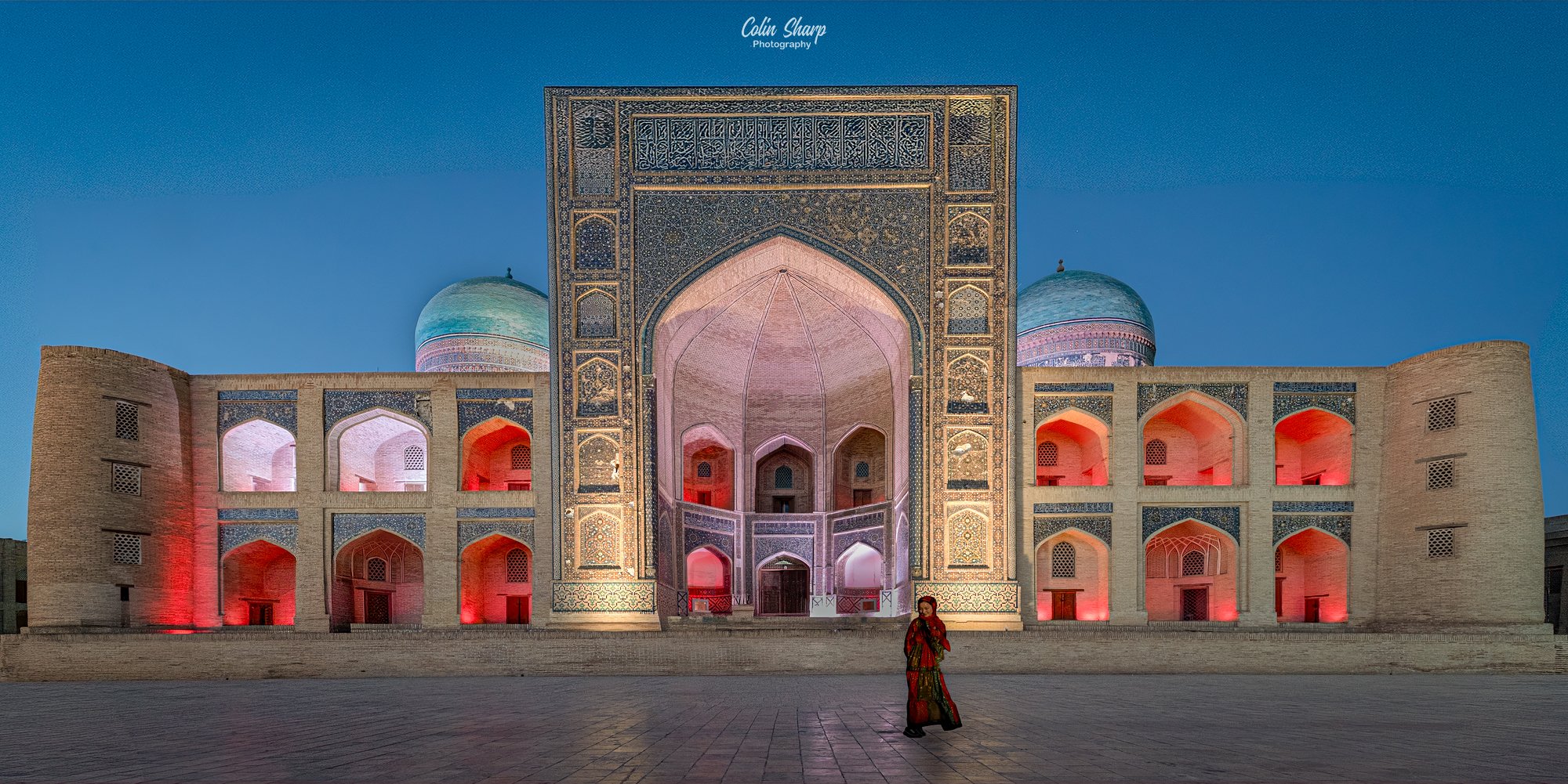 An evening blue hour photograph of the Mir-i Arab Medressa lit up by coloured floodlights with a locally dressed woman standing in the square in front of the Medressa.in front