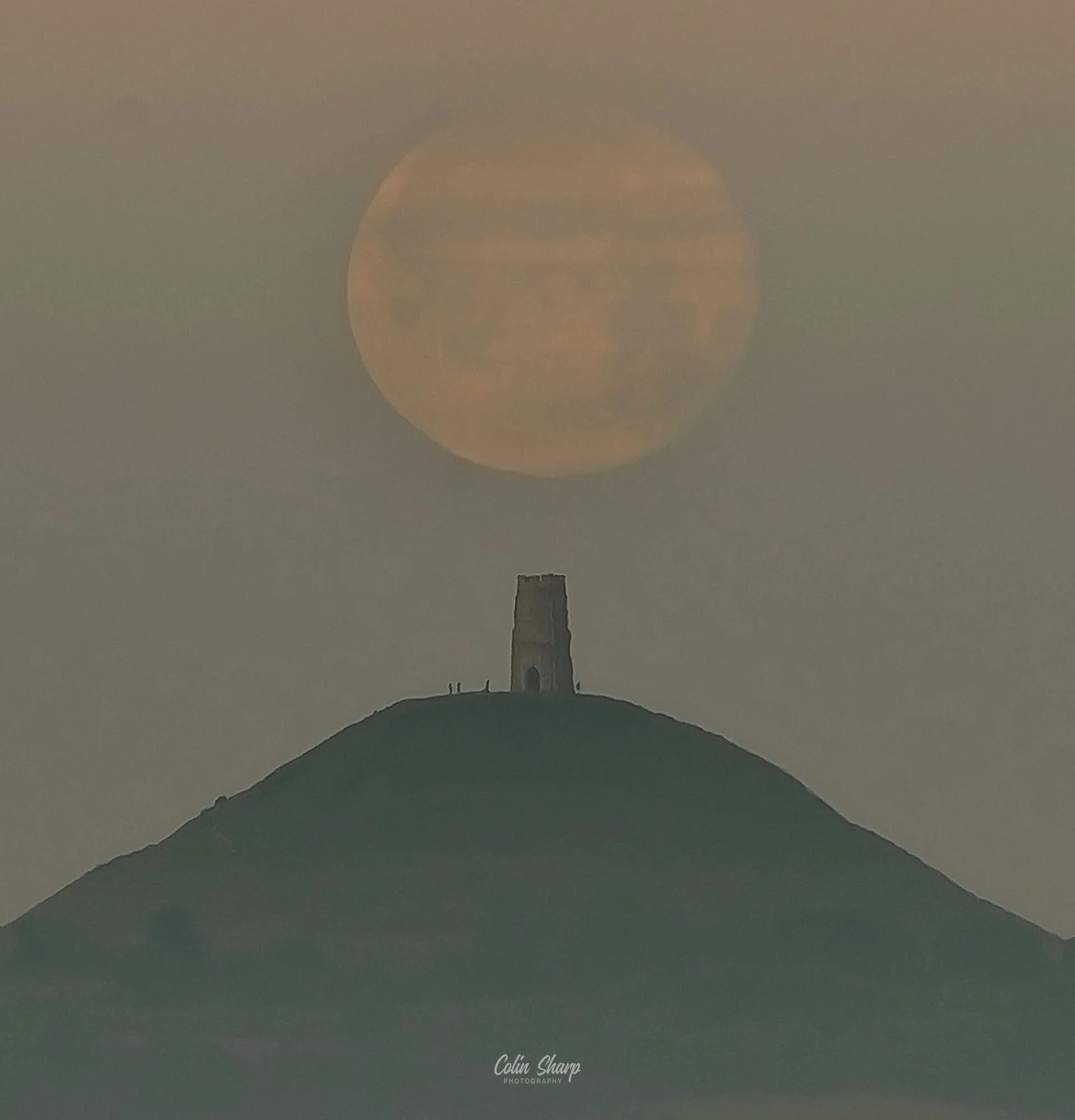 St Michaels Tower on Glastonbury Tor, a hilltop with an old stone tower under a large, orange tinted setting full super moon in the sky.