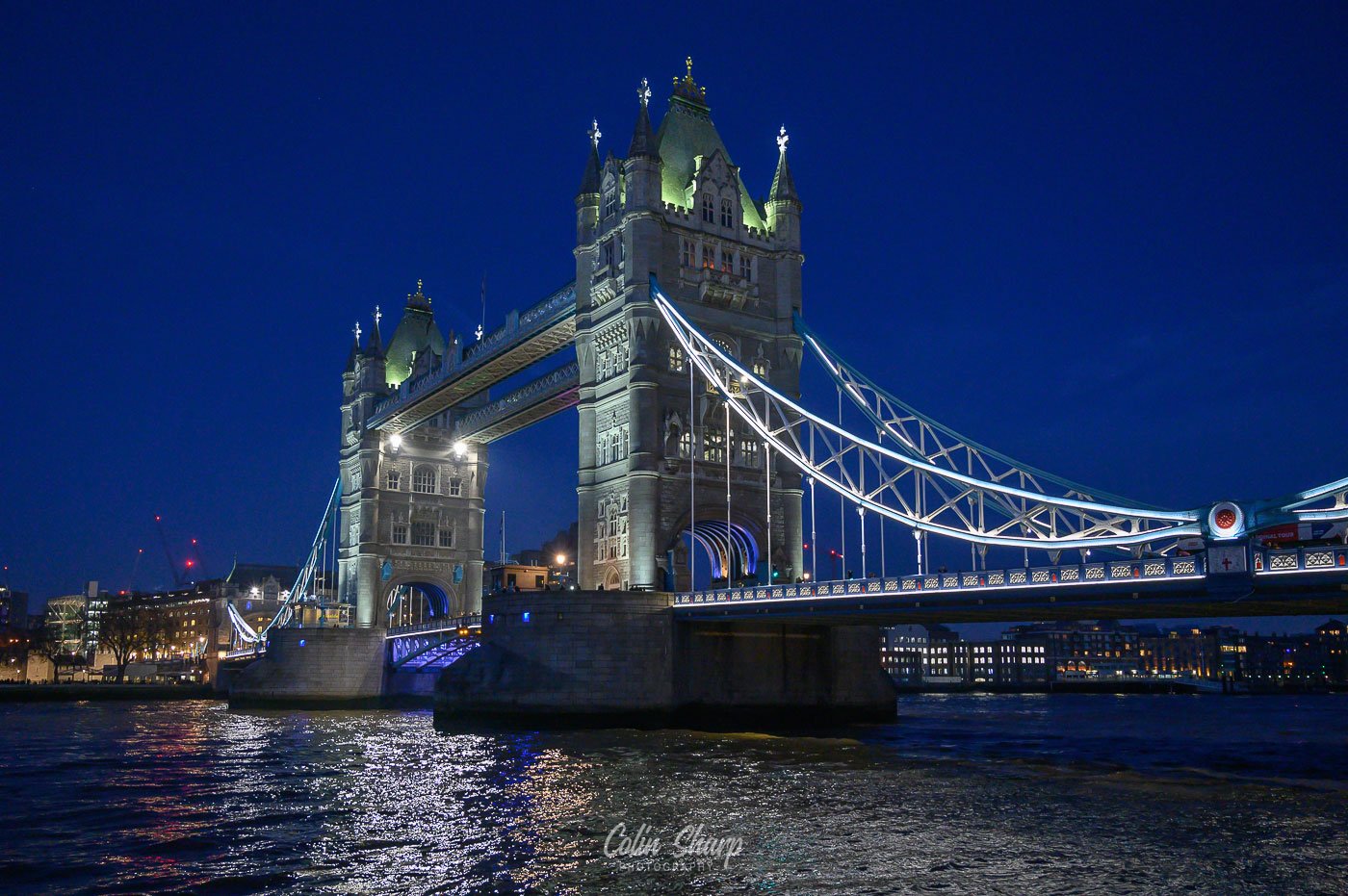 Late evening blue hour view of London's Tower Bridge illuminated with blue and white lights over the River Thames, city skyline in the background.