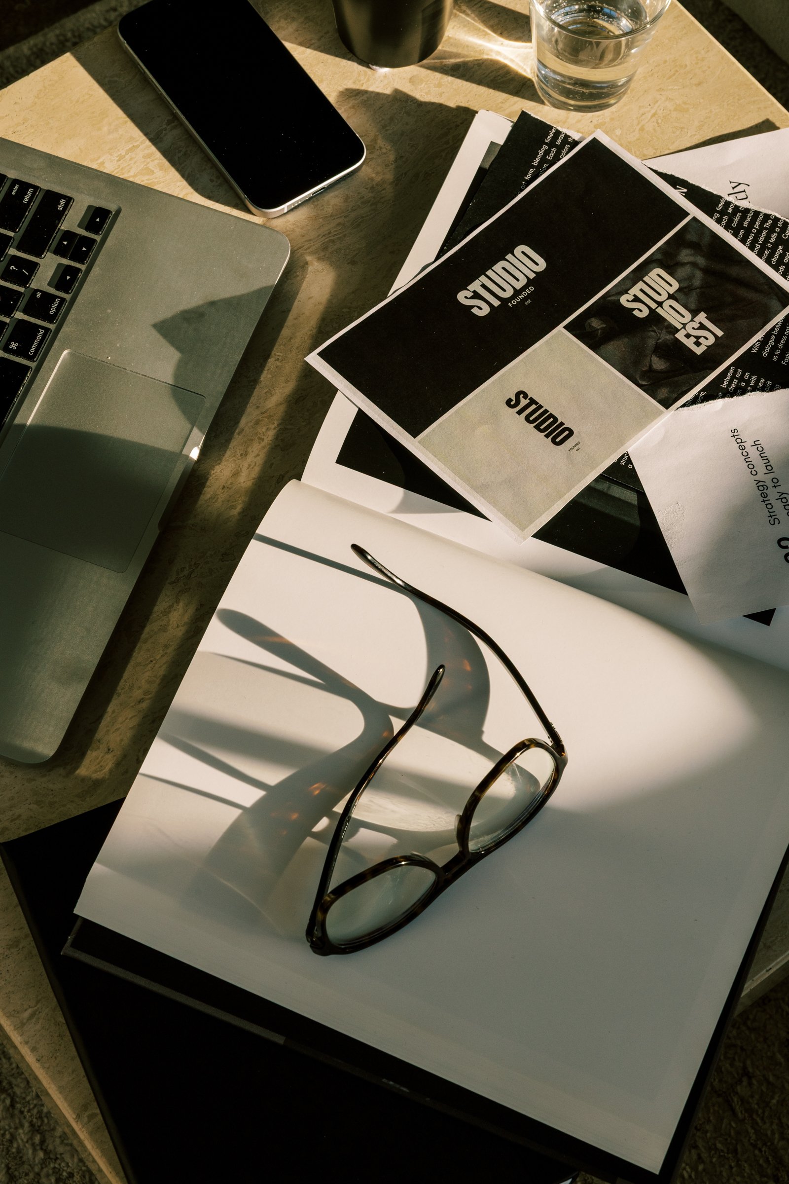 Eyeglasses sit on papers near a laptop, phone, and water glass on a desk.
