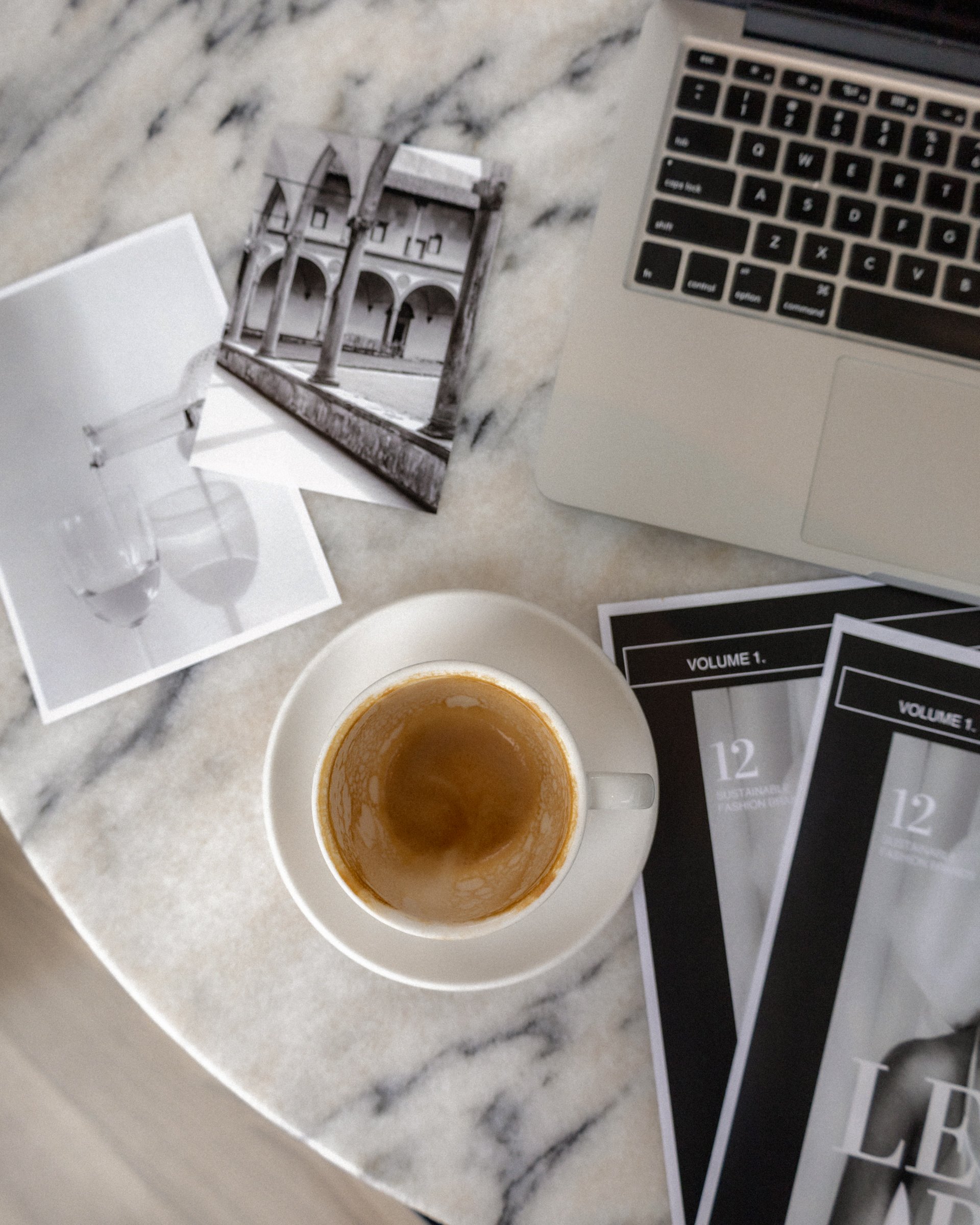 Flatlay of a coffee cup on a marble table with a laptop, magazines, and photos.