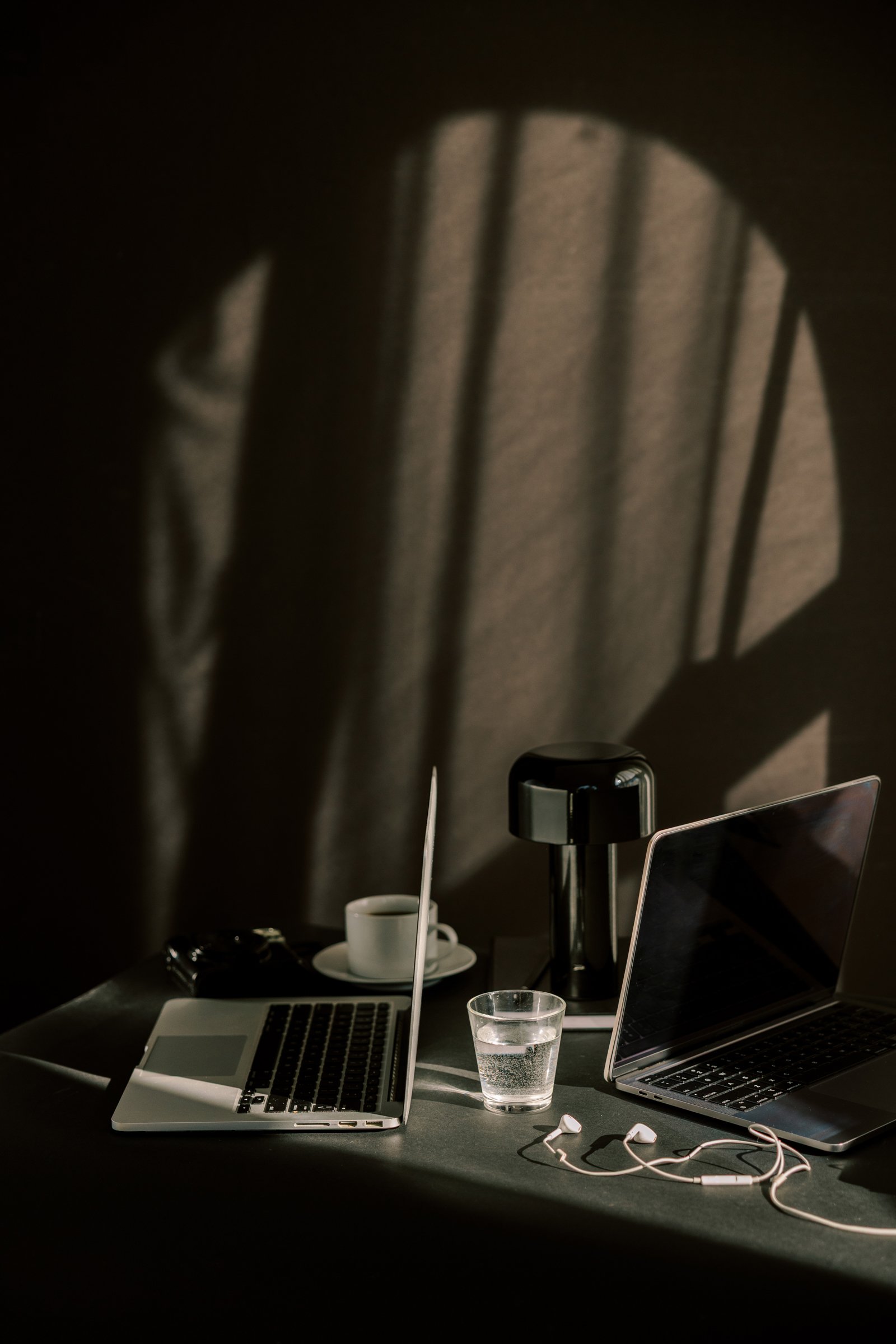 Two laptops and a coffee cup sit on a desk, bathed in moody light and shadow.