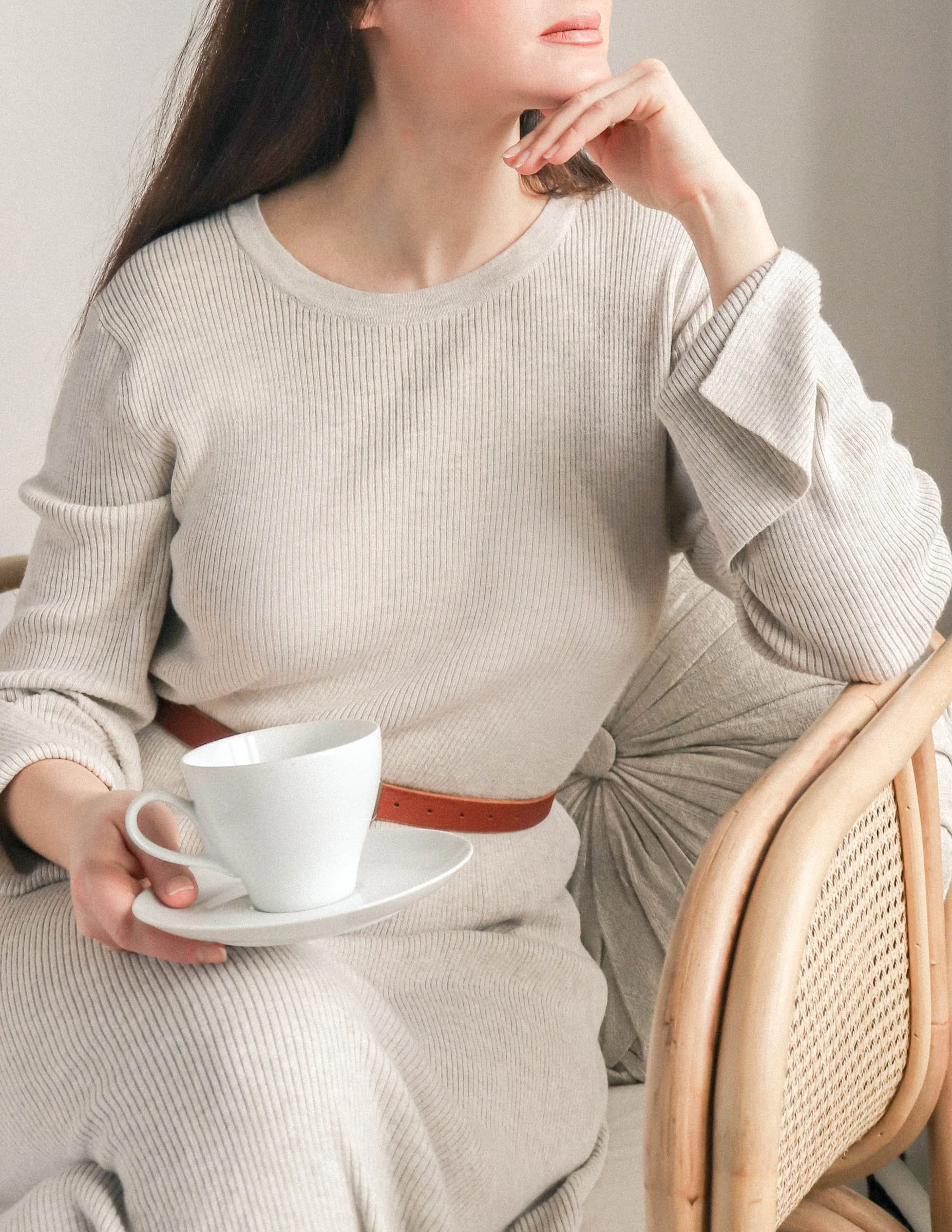 a woman wearing a beige dress with a brown belt around her center sitting in a chair holding a cup of coffee on a saucer. her hand is to her chin as she looks out the window