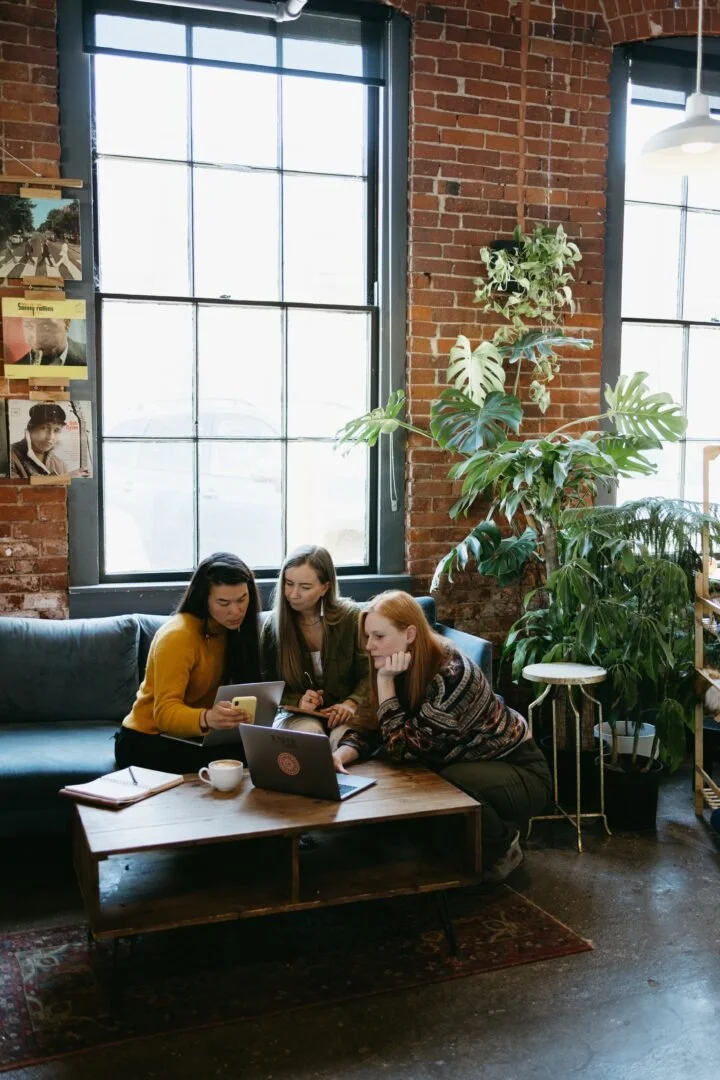 Three women working together over their laptops and cellphones in a coffee shop with a brick wall and monstera plant behind them