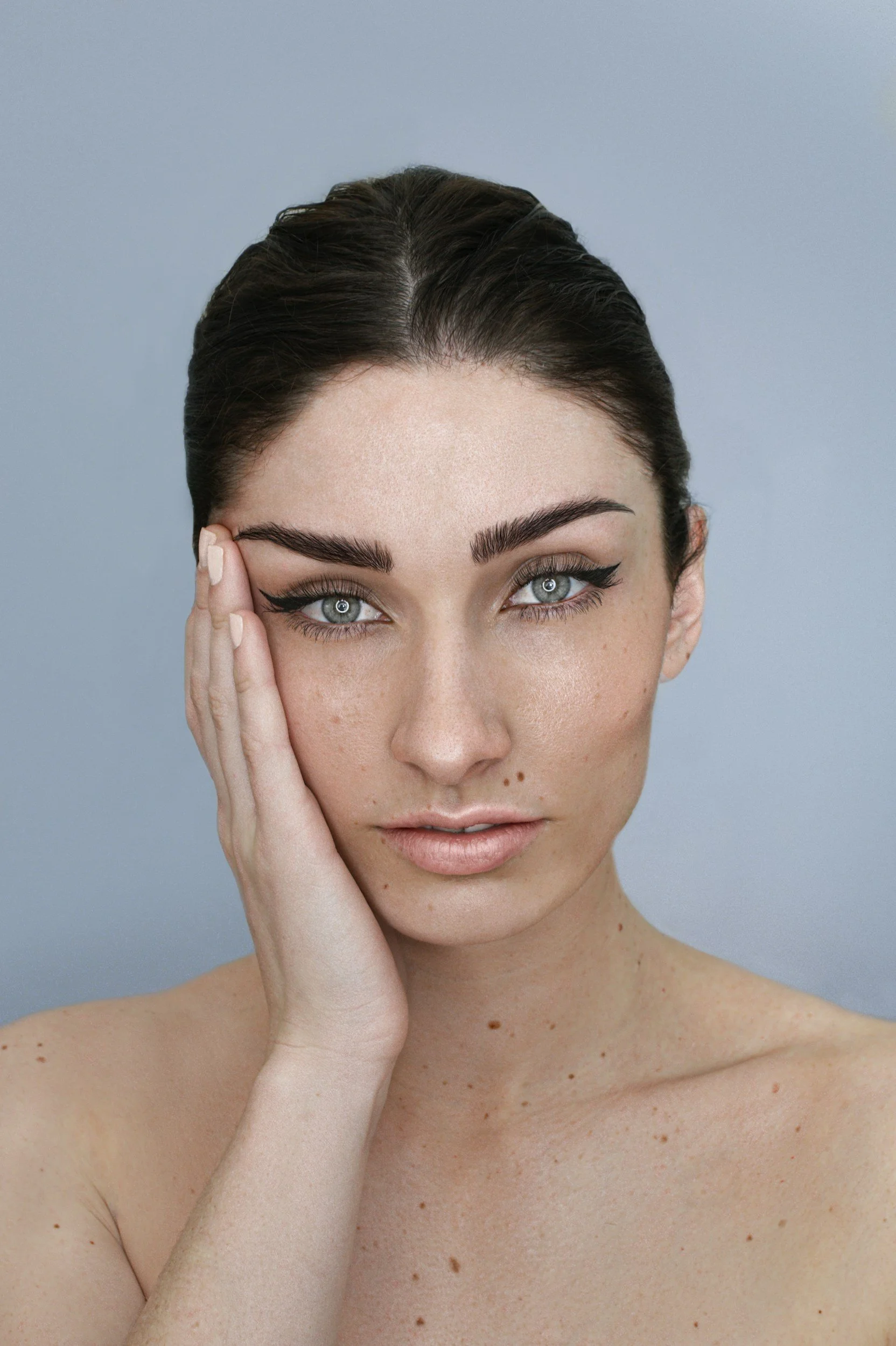 Close-up portrait of a young woman with dark hair, blue eyes, and natural freckles, touching her face gently, wearing light makeup, against a plain light blue background.