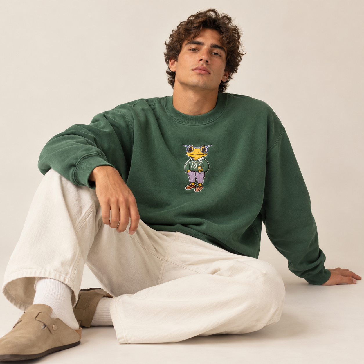 Man wearing Mr. Coquí forest green embroidered crewneck sweatshirt by 787 Coffee with cream wide-leg pants and tan clogs, relaxed oversized fit