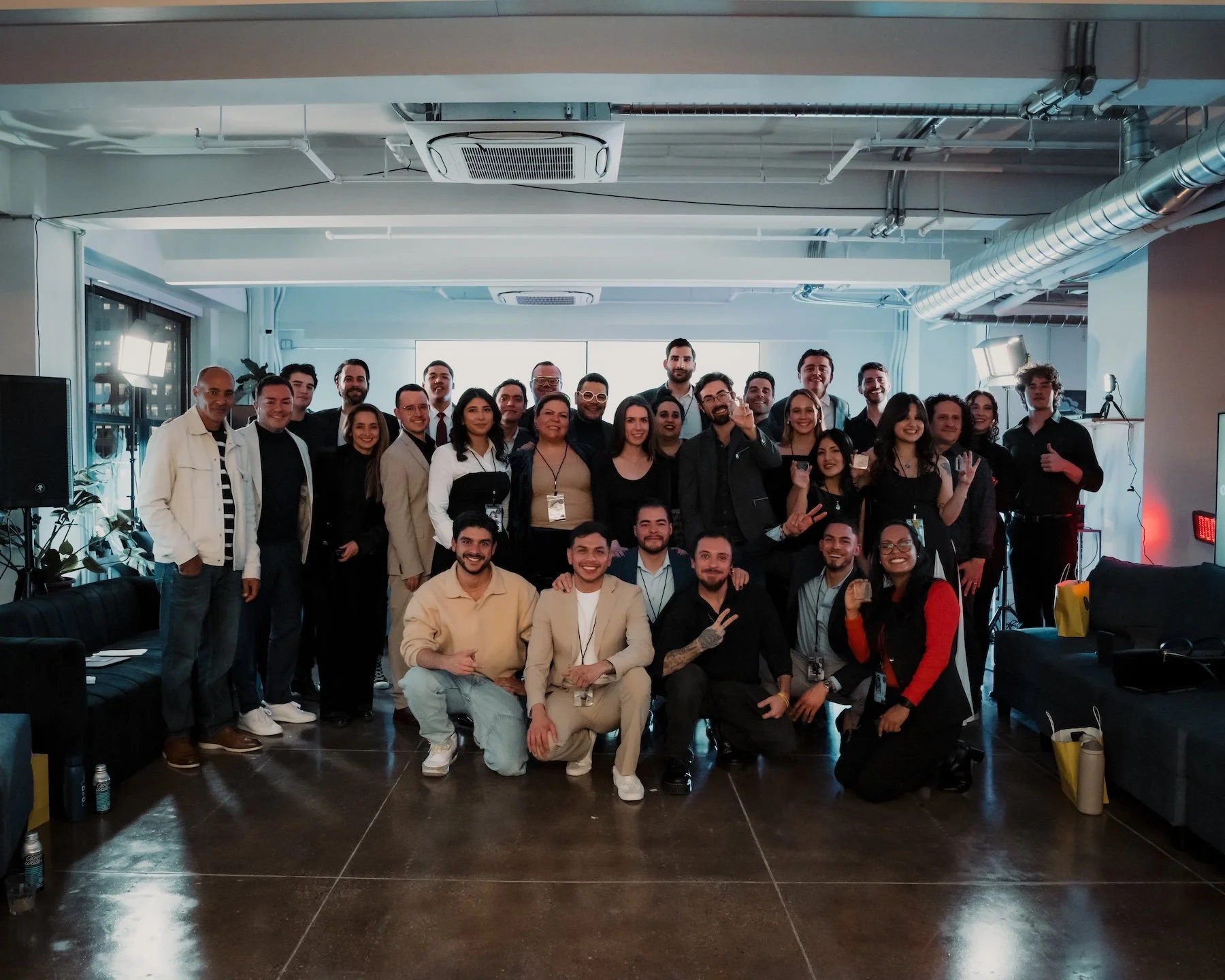 Group of diverse people posing for a photo in a modern office or conference room with bright natural and artificial lighting, some standing and others kneeling, smiling and making gestures.