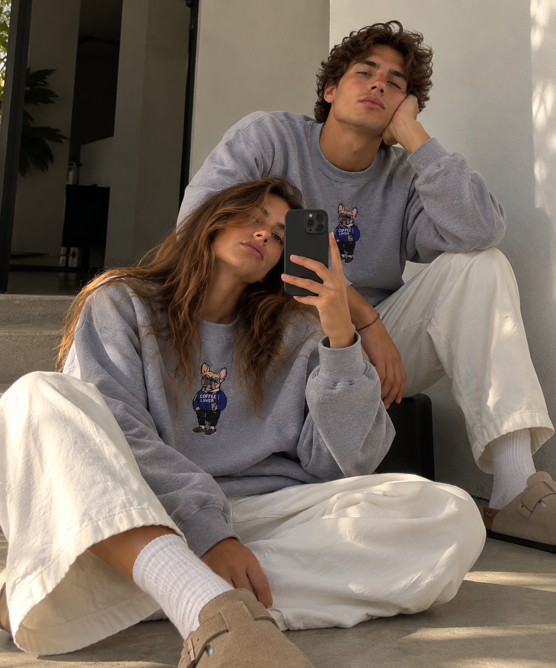 Man and woman wearing matching heather grey Frenchie Coffee Lover embroidered crewneck sweatshirts by 787 Coffee with cream wide-leg pants and suede clogs, relaxed oversized fit