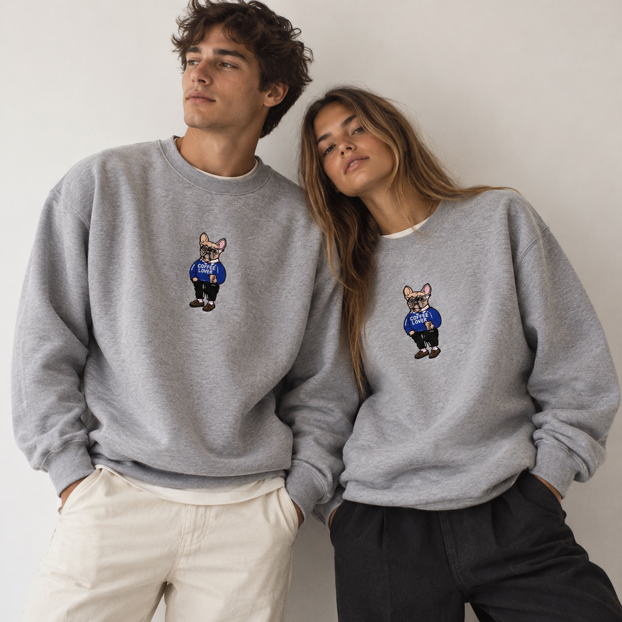 Man and woman wearing matching heather grey Frenchie Coffee Lover embroidered crewneck sweatshirts by 787 Coffee, relaxed unisex fit, shown with cream and black pants