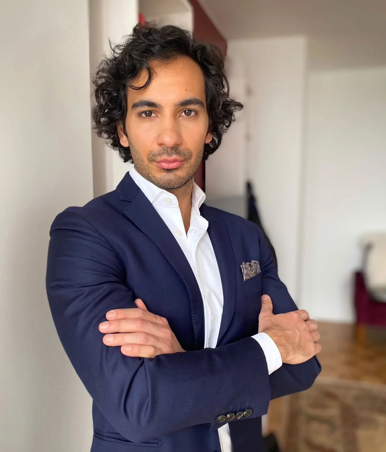 A man with dark, curly hair and light brown skin, wearing a navy blue suit jacket with a white dress shirt underneath, standing with arms crossed in a room with white walls and wood flooring.