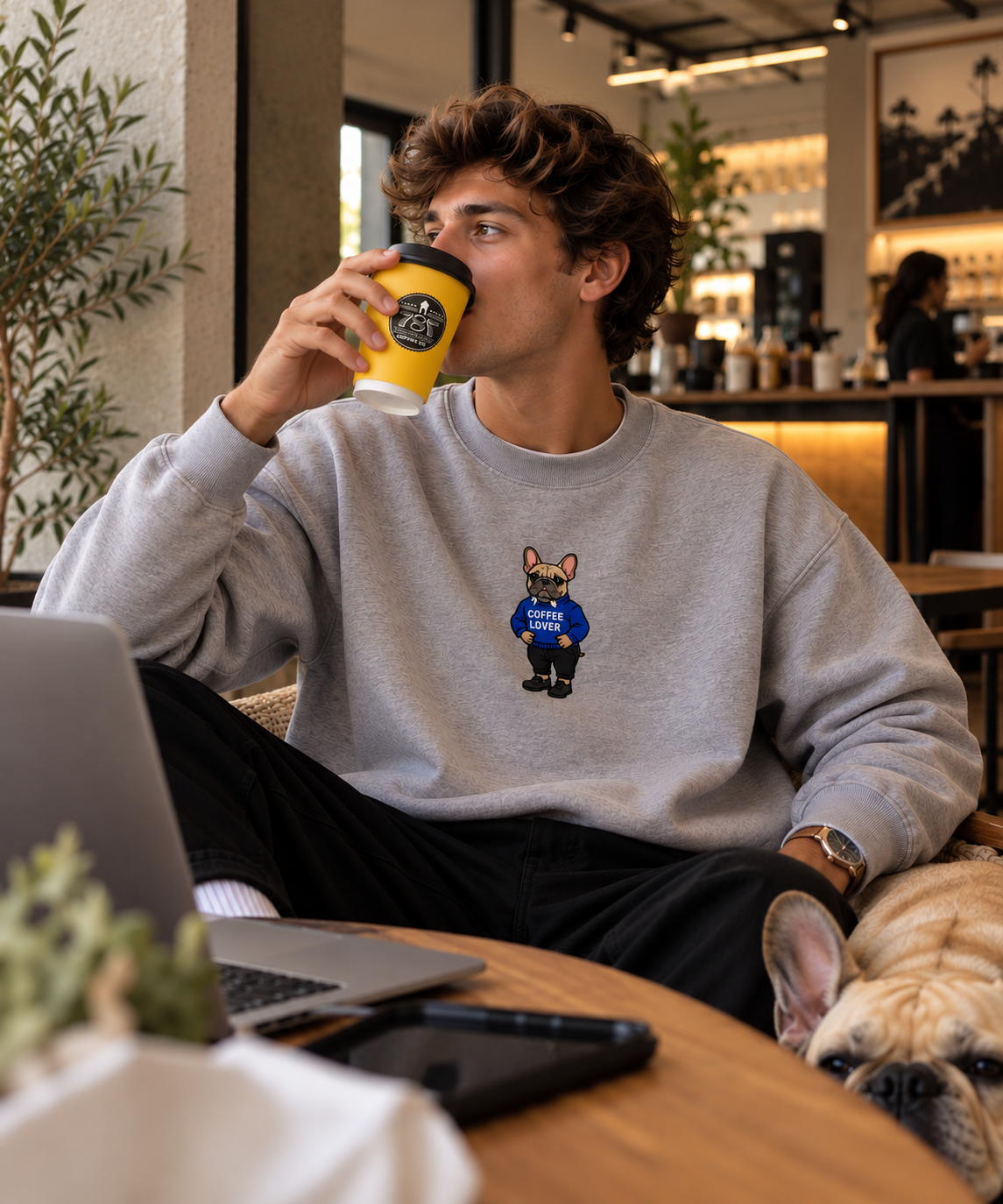 Man drinking from a yellow 787 Coffee cup at a café table with a laptop, wearing a heather grey Frenchie Coffee Lover embroidered crewneck sweatshirt, a real French bulldog resting beside him