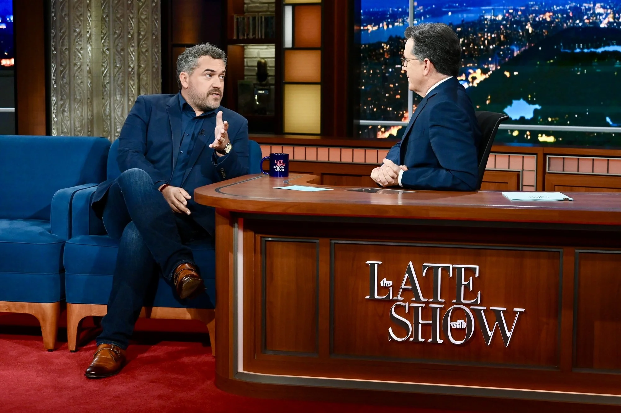 TV talk show set featuring guest and host men in conversation. The guest has curly hair, beard, navy blazer, and jeans, sitting on blue couch. The host has dark hair, glasses, navy suit, sitting behind wooden desk with show logo. Background shows cityscape at night with lights.