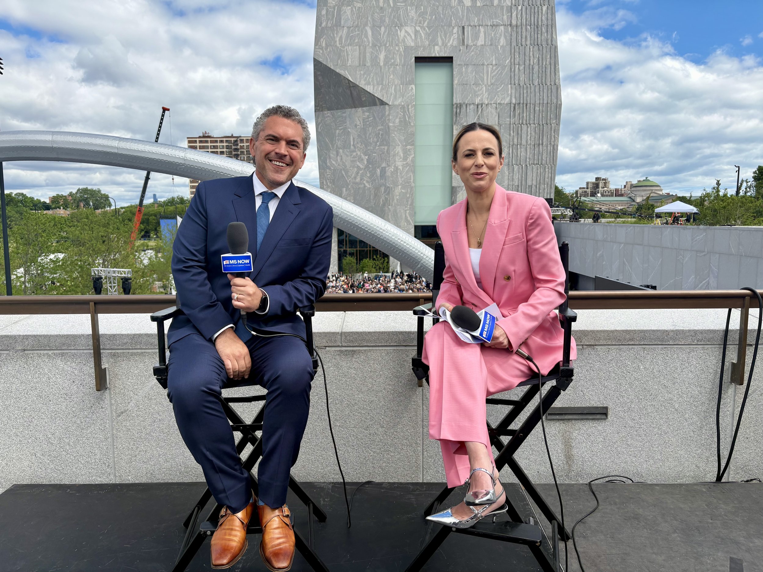 Two news anchors sitting on black foldable chairs outdoors, smiling at the camera, with microphones in front of them, a modern building and a cloudy sky behind them.