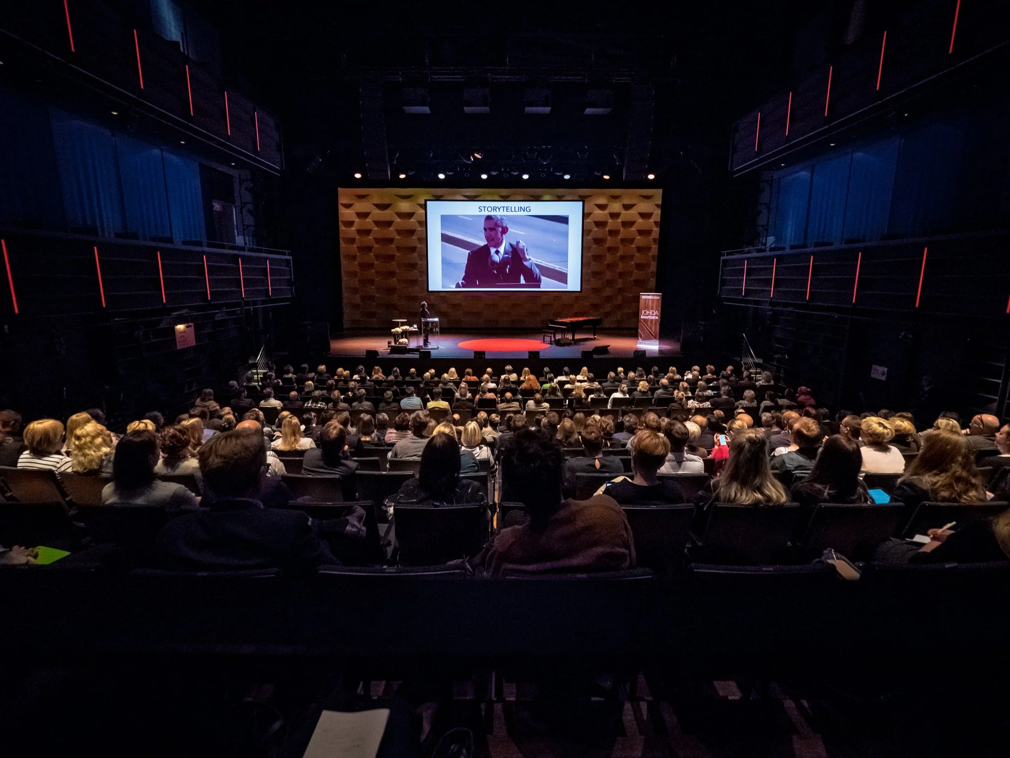 A large audience seated in a theater watching a presentation on stage. The presentation slide shows a man in a suit with the word 'STORYTELLING' at the top.