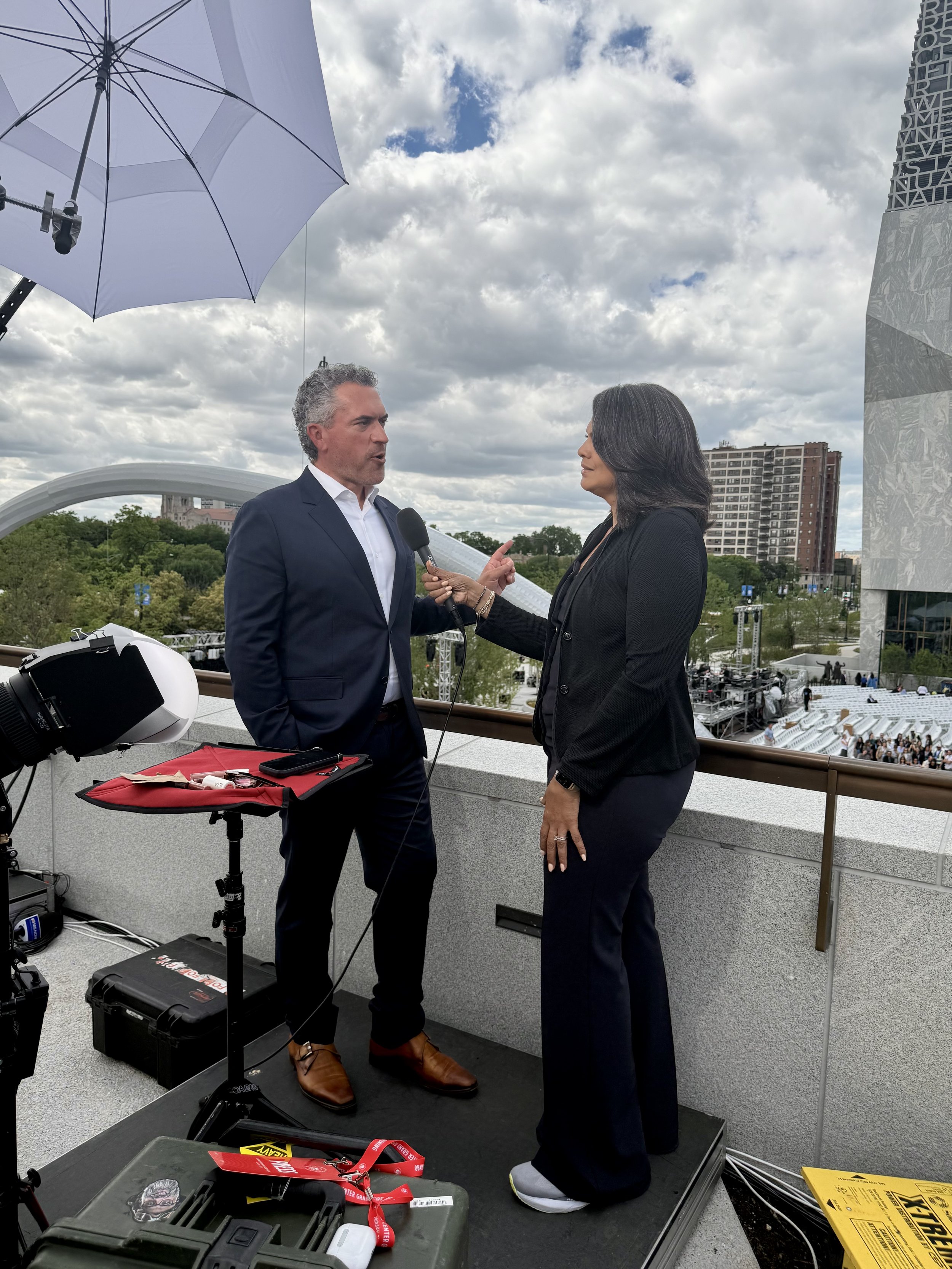 A man in a suit and a woman in a blazer are engaged in an interview on an outdoor rooftop with a cloudy sky. The man is holding a microphone and appears to be speaking, while the woman listens attentively. There is camera equipment, cables, and a helmet on a table nearby. In the background, there are trees, a modern building, and an audience seating area.