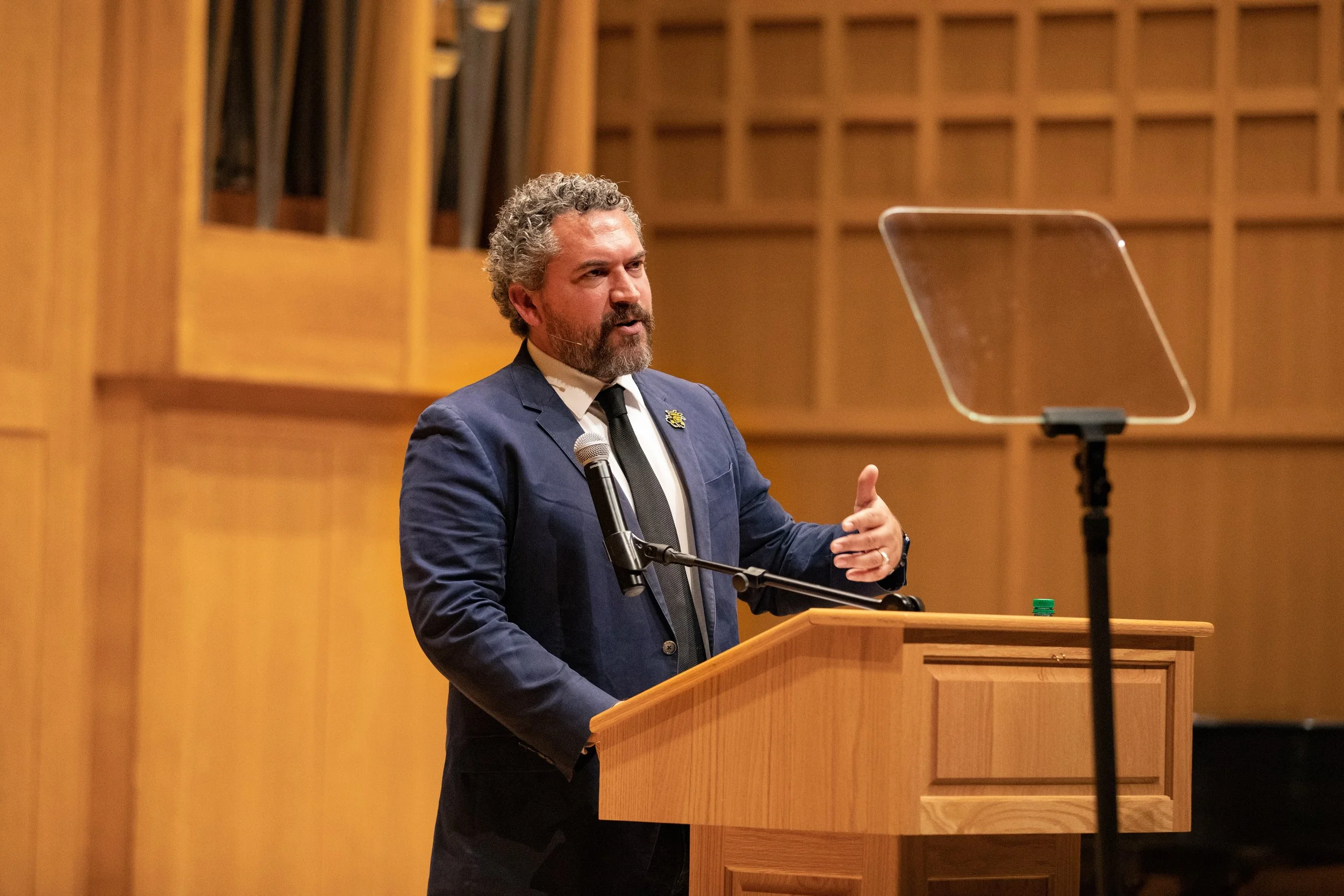 A man with curly gray hair and a beard, dressed in a dark blue suit, white shirt, and black tie, speaking at a podium in a wood-paneled room. There is a microphone attached to his suit and a teleprompter in front of him.